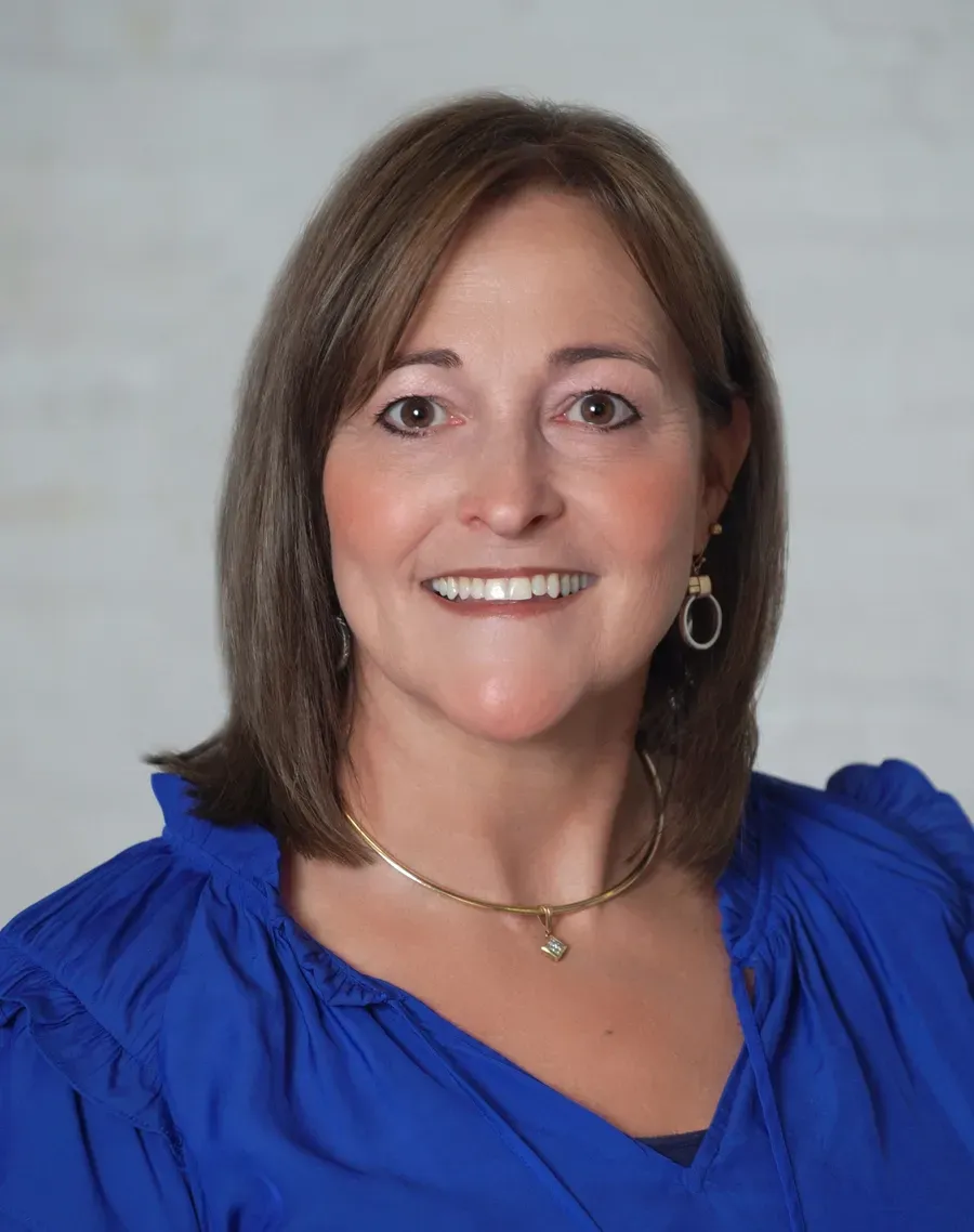 Woman with light brown hair, smiling, wearing a blue blouse and gold jewelry.