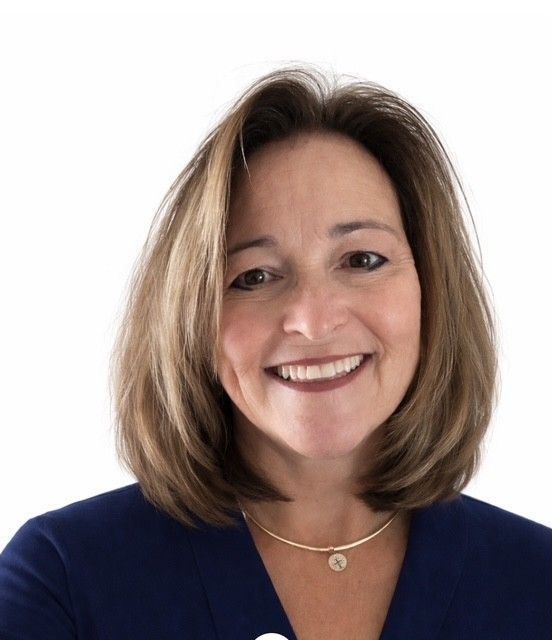 Woman with light brown hair, smiling, wearing a blue blouse and gold jewelry.