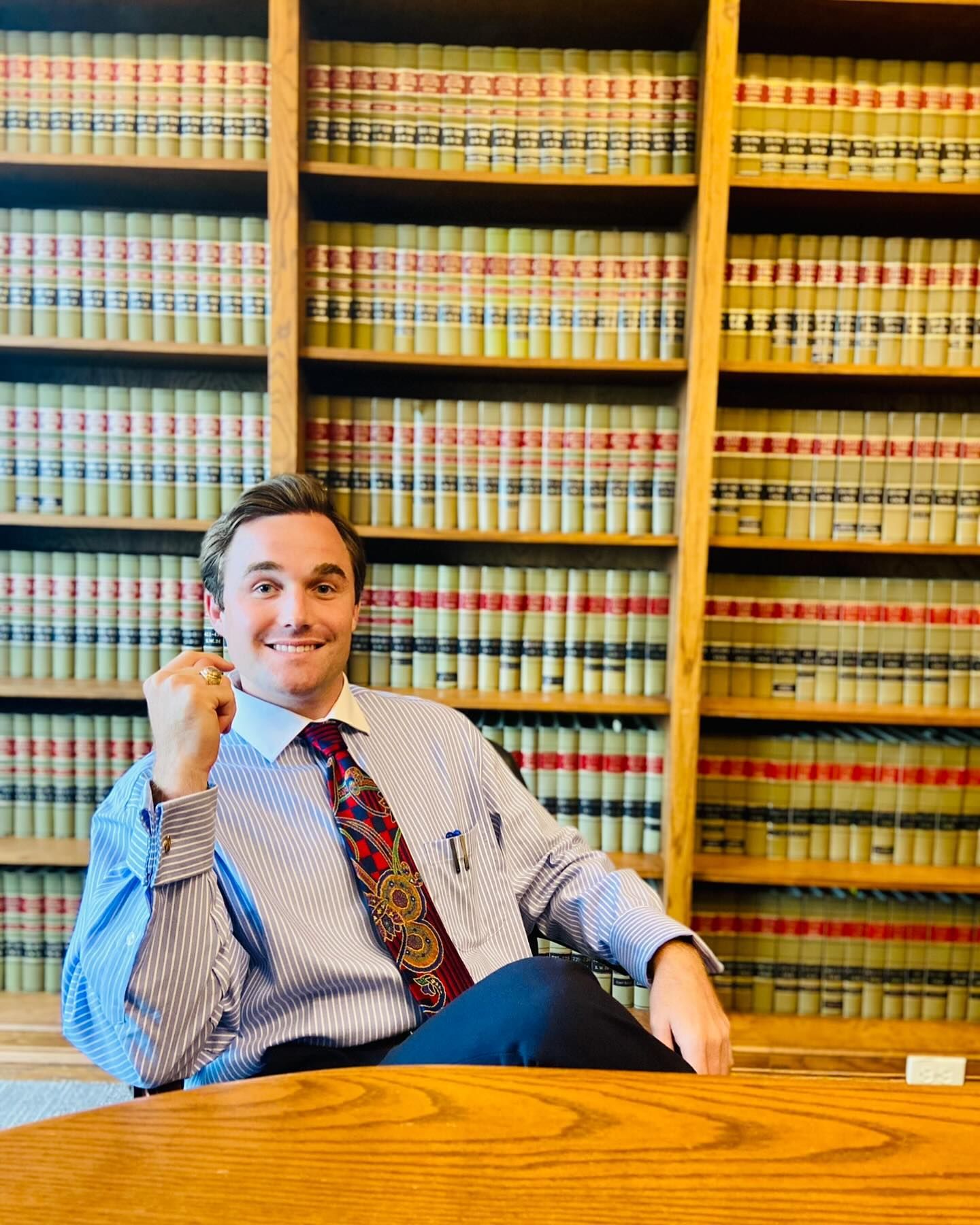Man smiles, fist raised, sitting in a law library. He wears a blue-striped shirt and tie. Bookshelves fill the background.