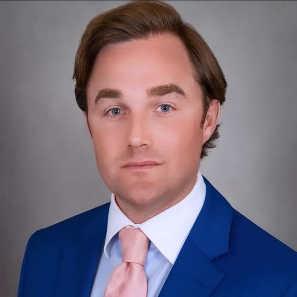 Man in a blue suit and pink tie, looking at the camera, against a gray backdrop.