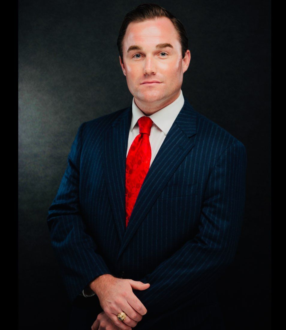Man in a pinstripe suit and red tie, arms crossed, posing against a dark background.