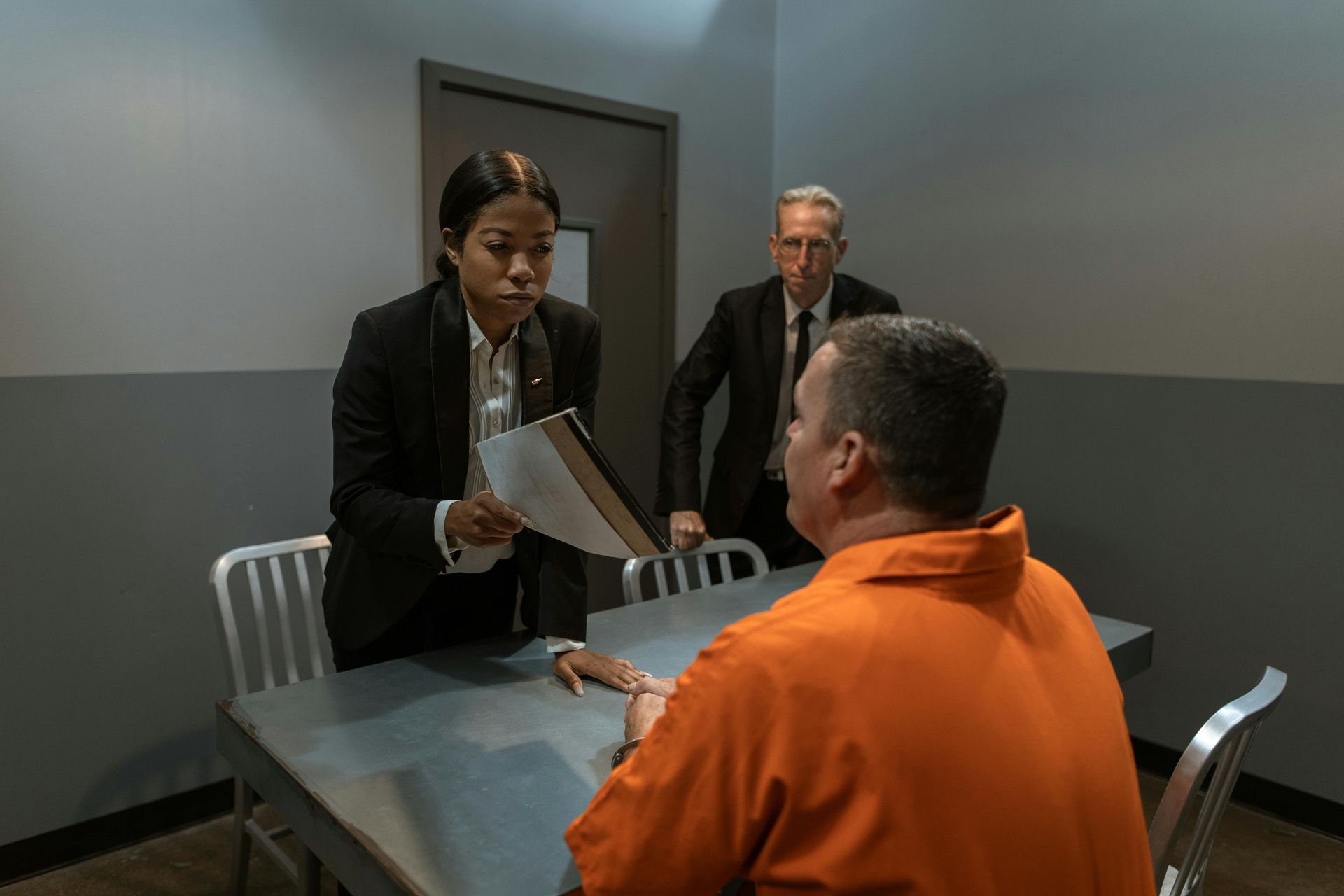 A woman in a suit shows papers to a man in orange jail clothes, with a man in a suit standing behind.