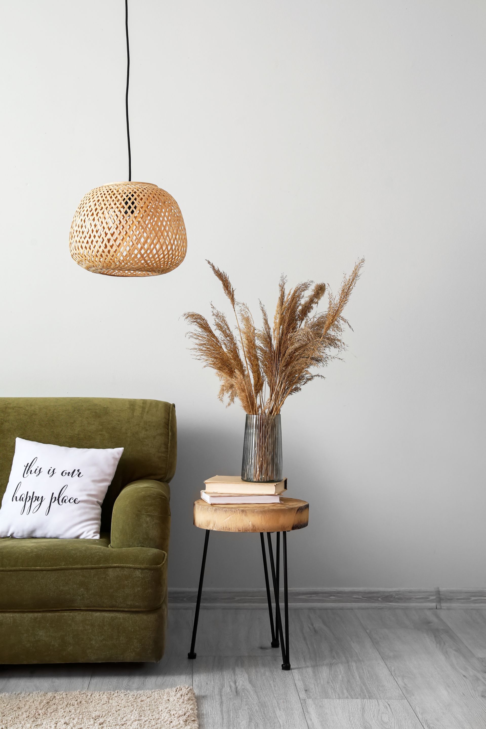 A green armchair, a small wood side table with dried grass in a vase and books, and a woven pendant light in a room.