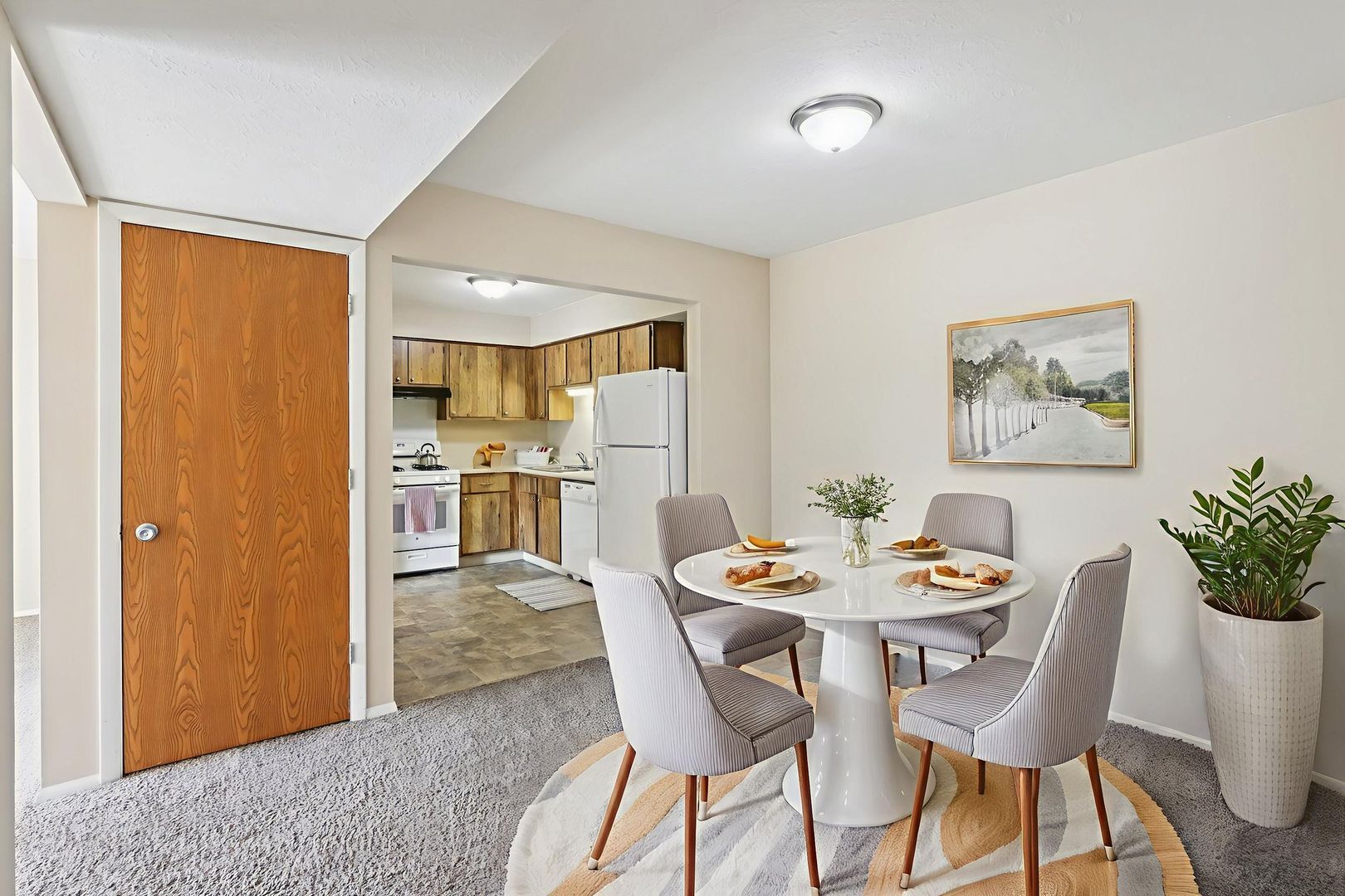 A dining area with a round white table, four gray chairs, and a potted plant, leading to a kitchen with wooden cabinets.