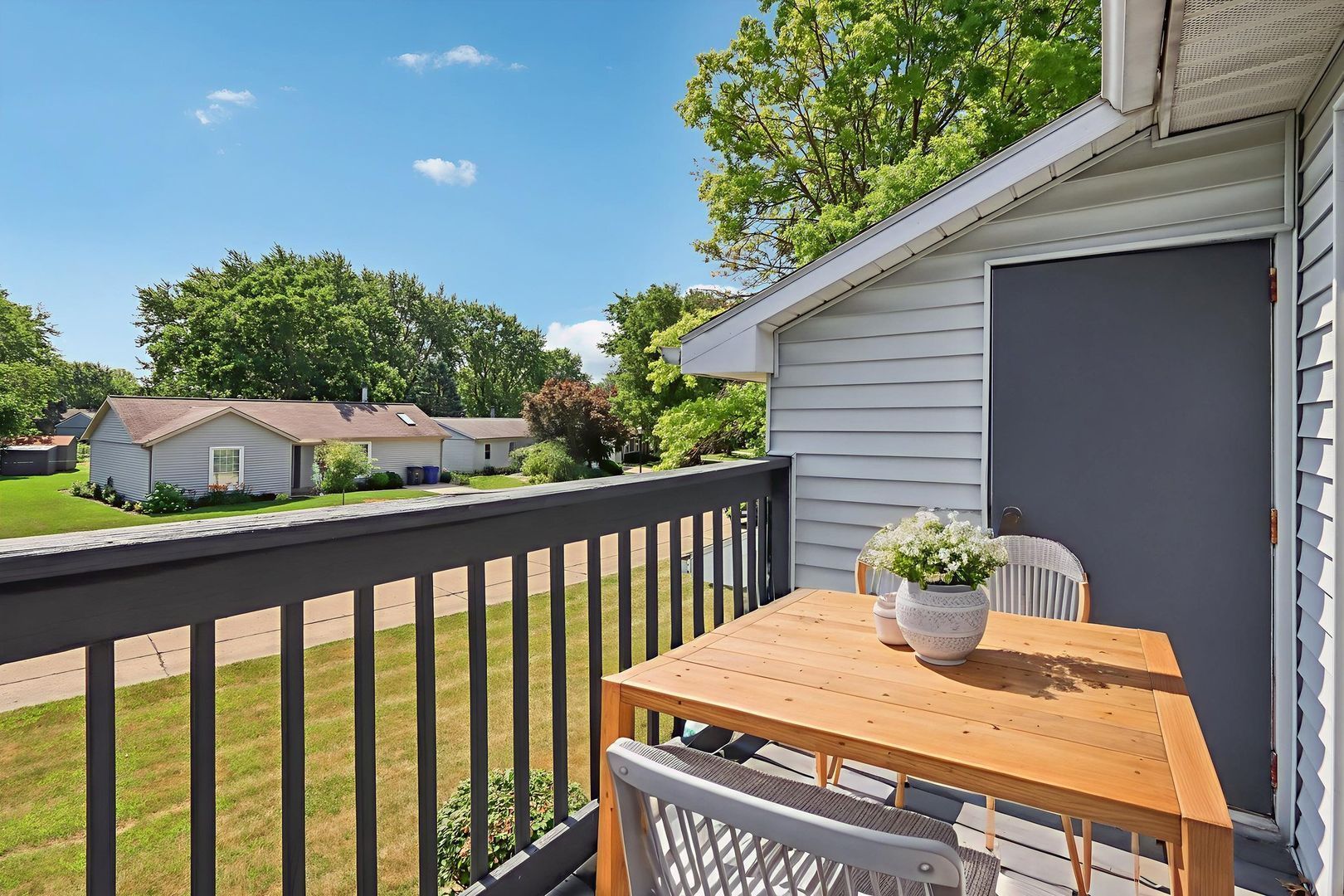 A small wooden table and two chairs on an outdoor balcony overlooking a grassy neighborhood on a sunny day.