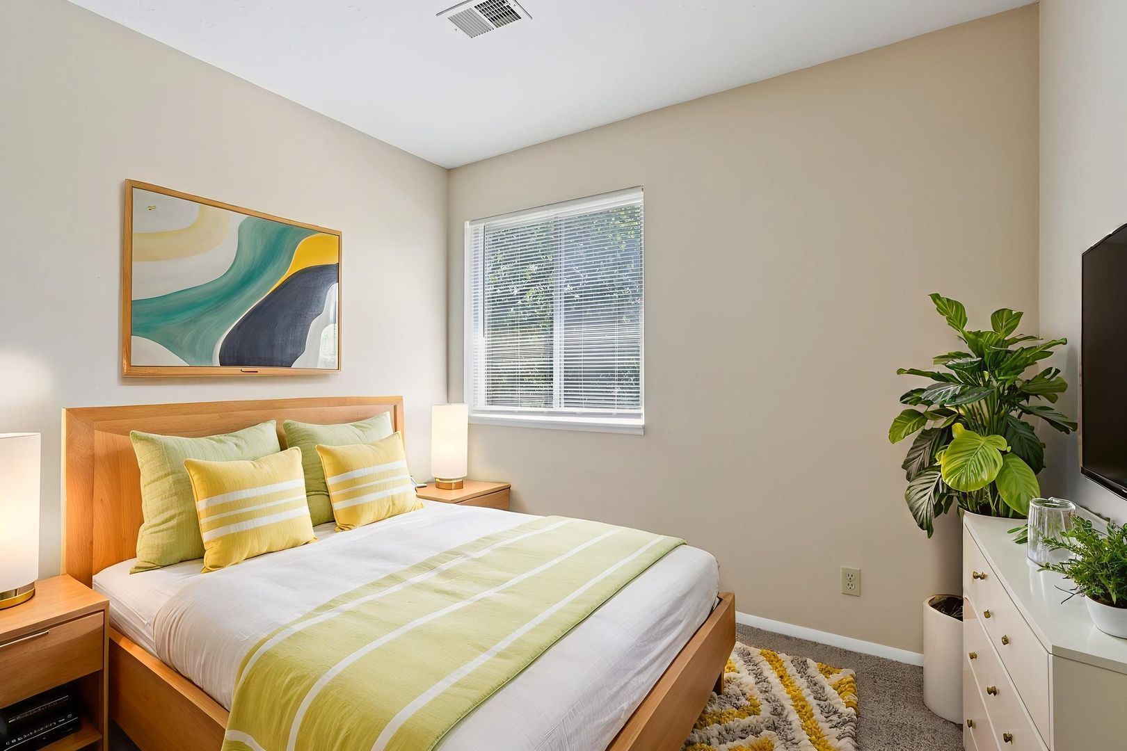 A bright bedroom with a double bed featuring yellow accents, a window with blinds, a potted plant, and wall art.