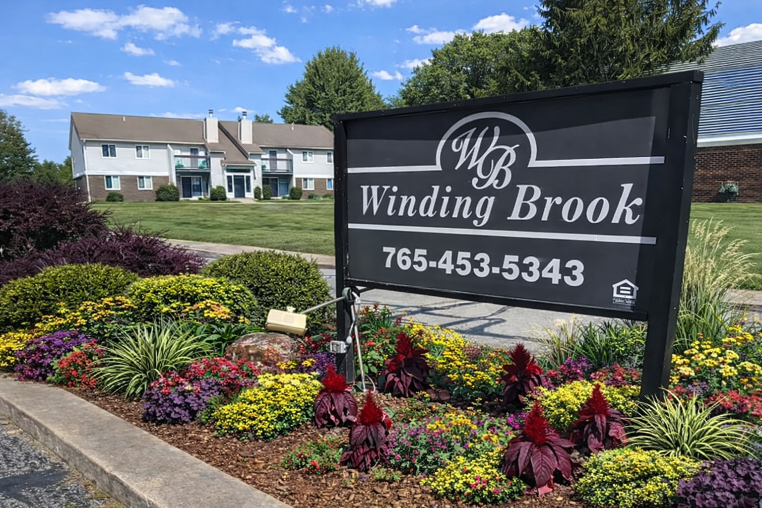 A Winding Brook apartment sign stands in a flowerbed in front of a residential building under a blue, sunny sky.