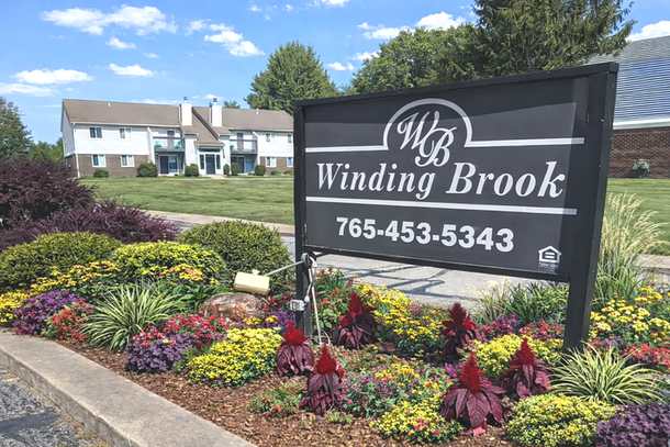 A black sign for Winding Brook apartments with phone number 765-453-5343, surrounded by a colorful garden, outside buildings.