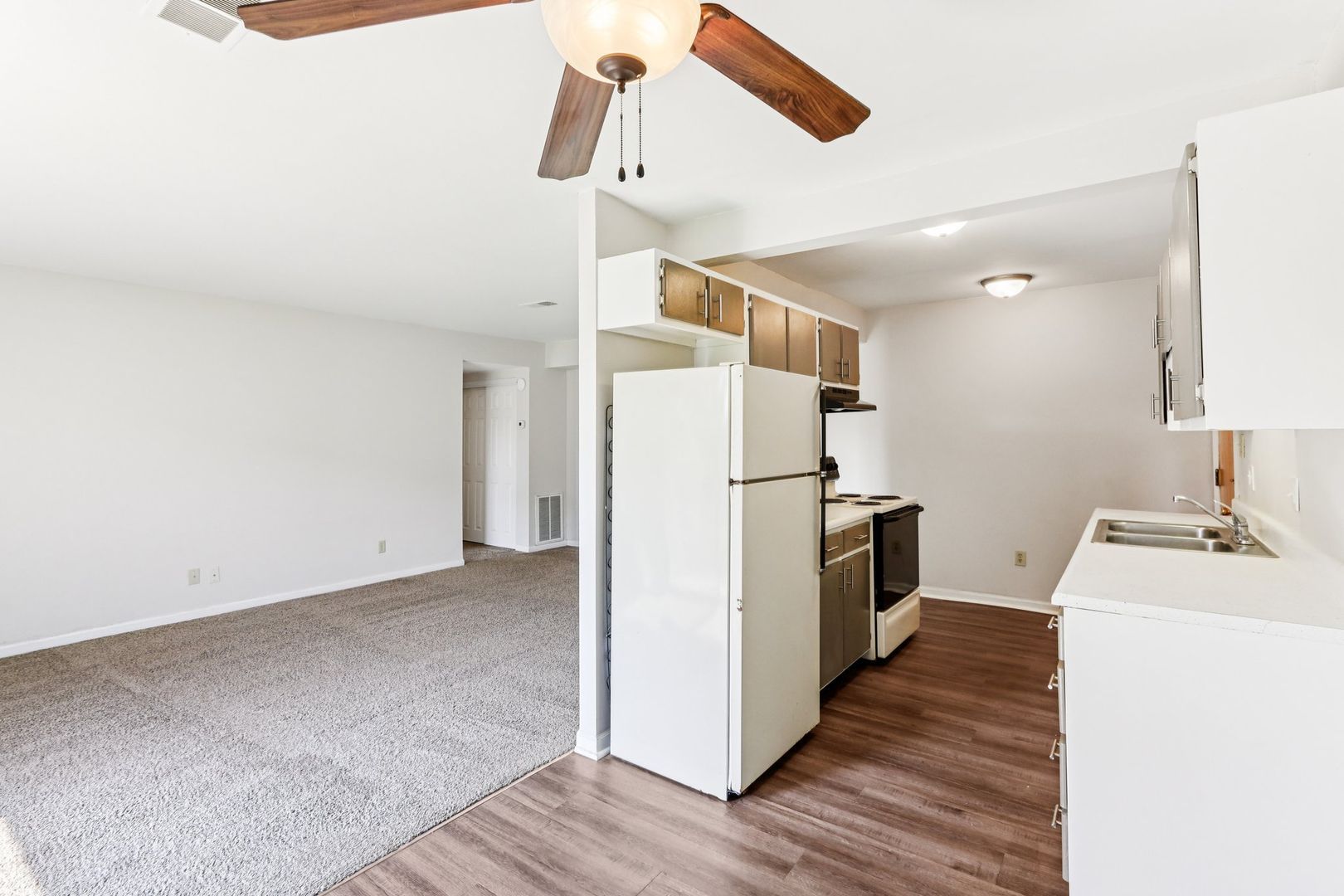 An open-concept apartment with a carpeted living room and a kitchen featuring white cabinets, appliances, and wood floors.