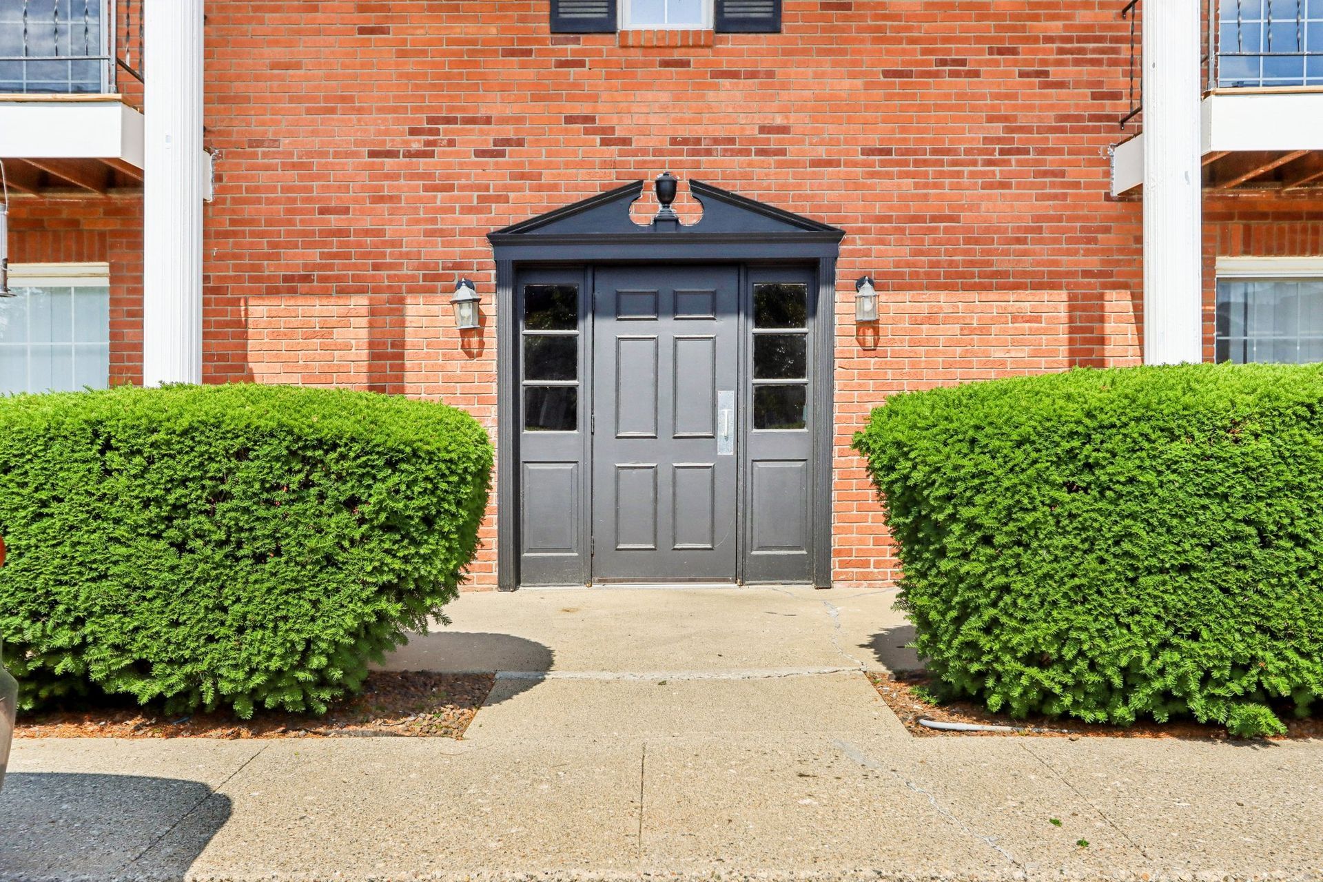 A dark grey double door with a pediment sits centered in a brick building exterior, flanked by two large, round bushes.