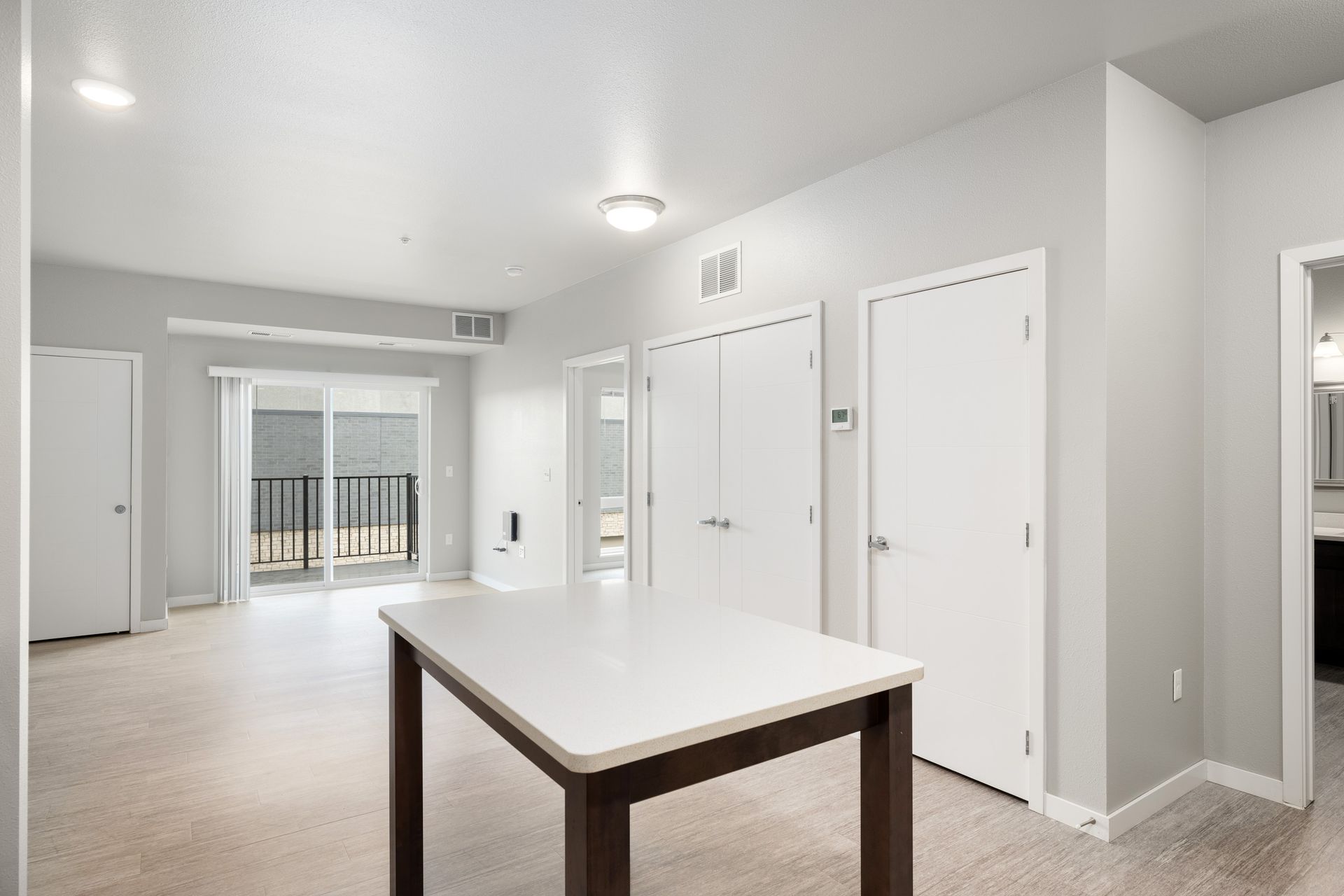 Spacious, bright apartment interior with a white table, balcony, and closed doors.