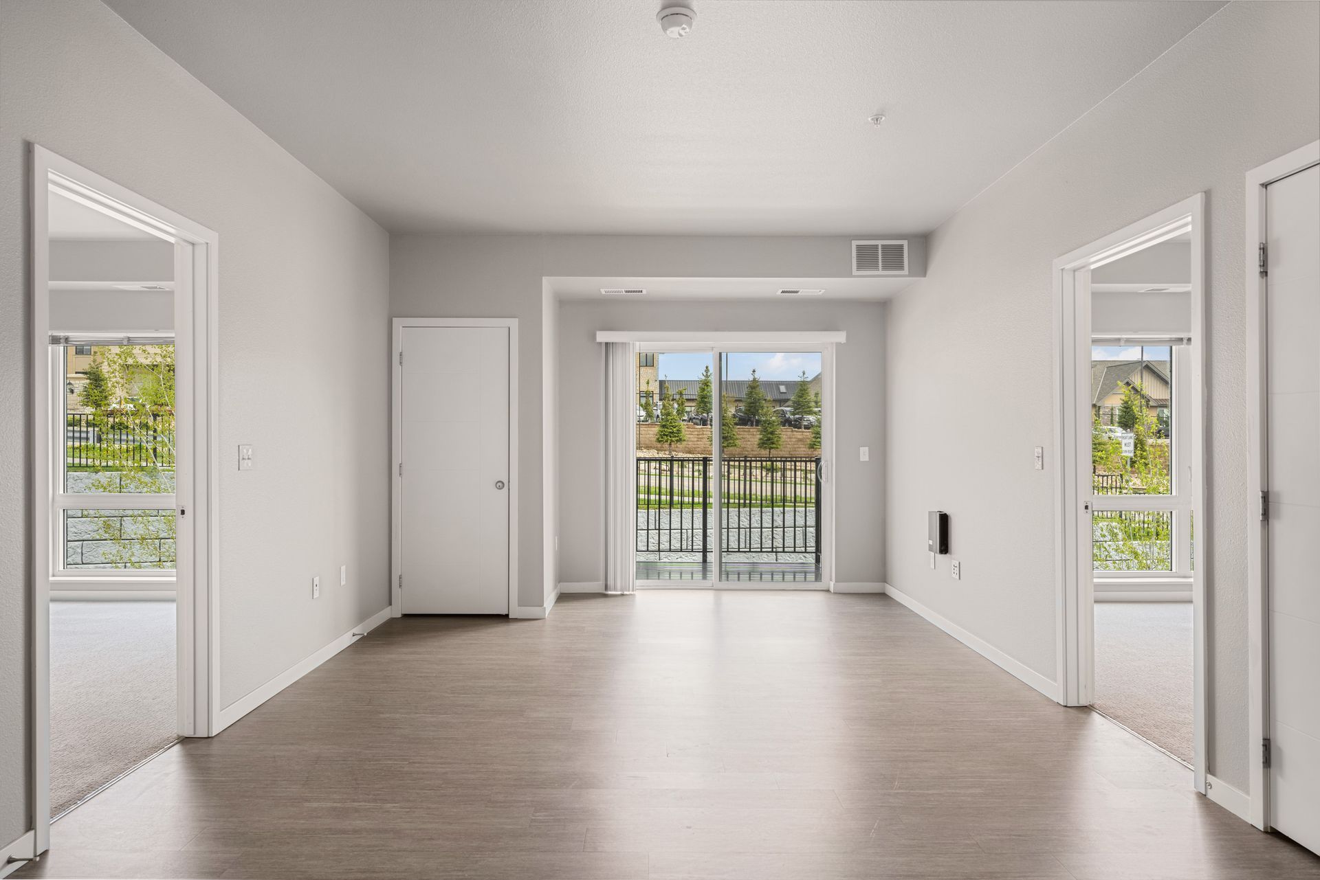 Empty, bright room with laminate flooring, doors, and a balcony. Windows offer a view of greenery.