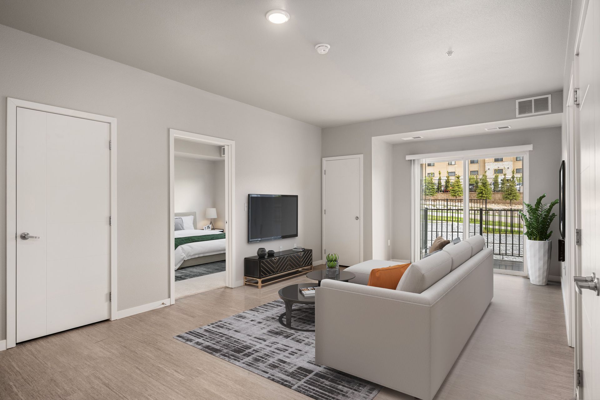 Modern living room with a couch, TV, and balcony access. Light gray walls, light wood floors.