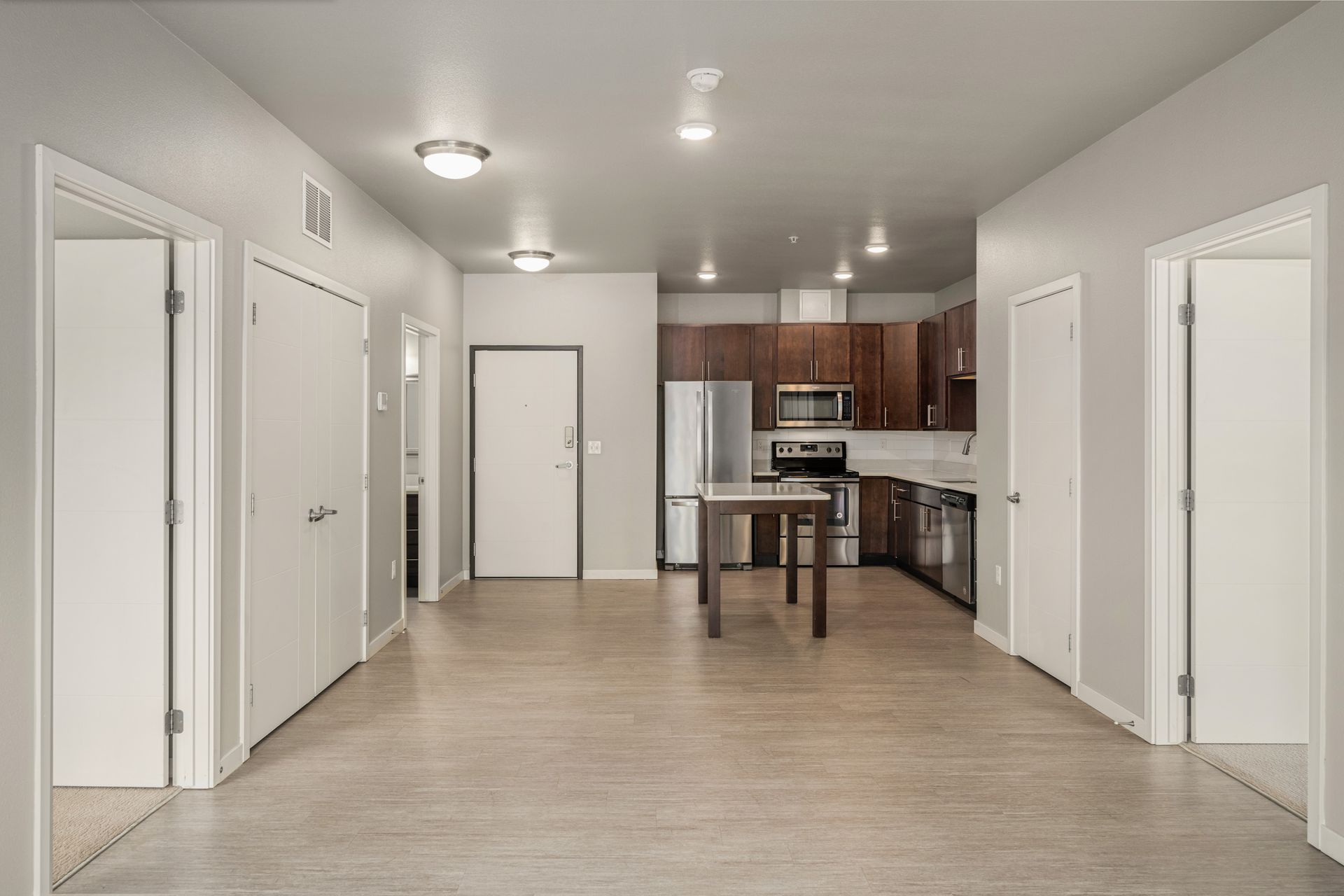 Empty apartment interior with kitchen, wooden floor, and several white doors.