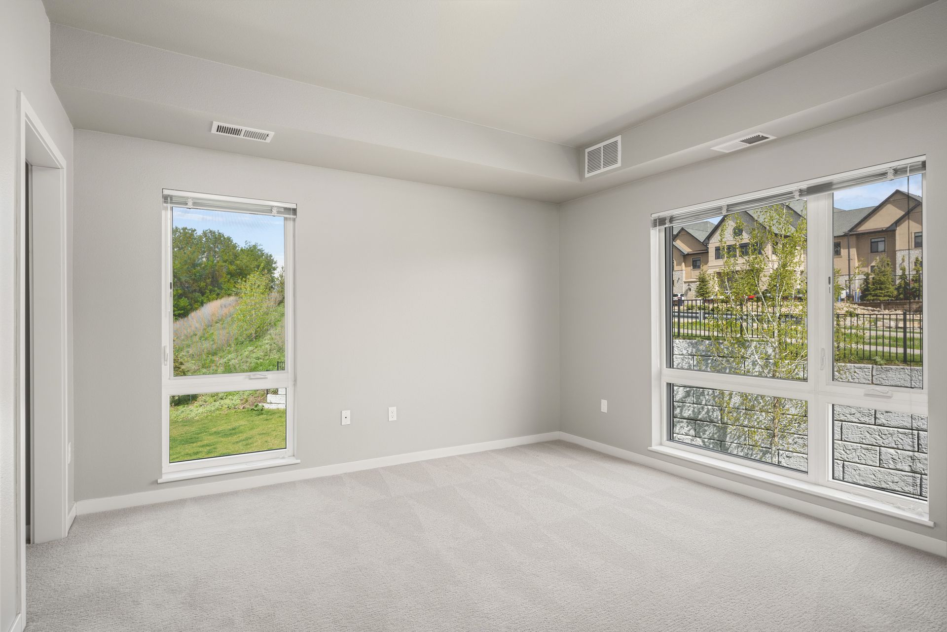 Empty room with two windows showing a green lawn and houses, with a light gray carpet and walls.