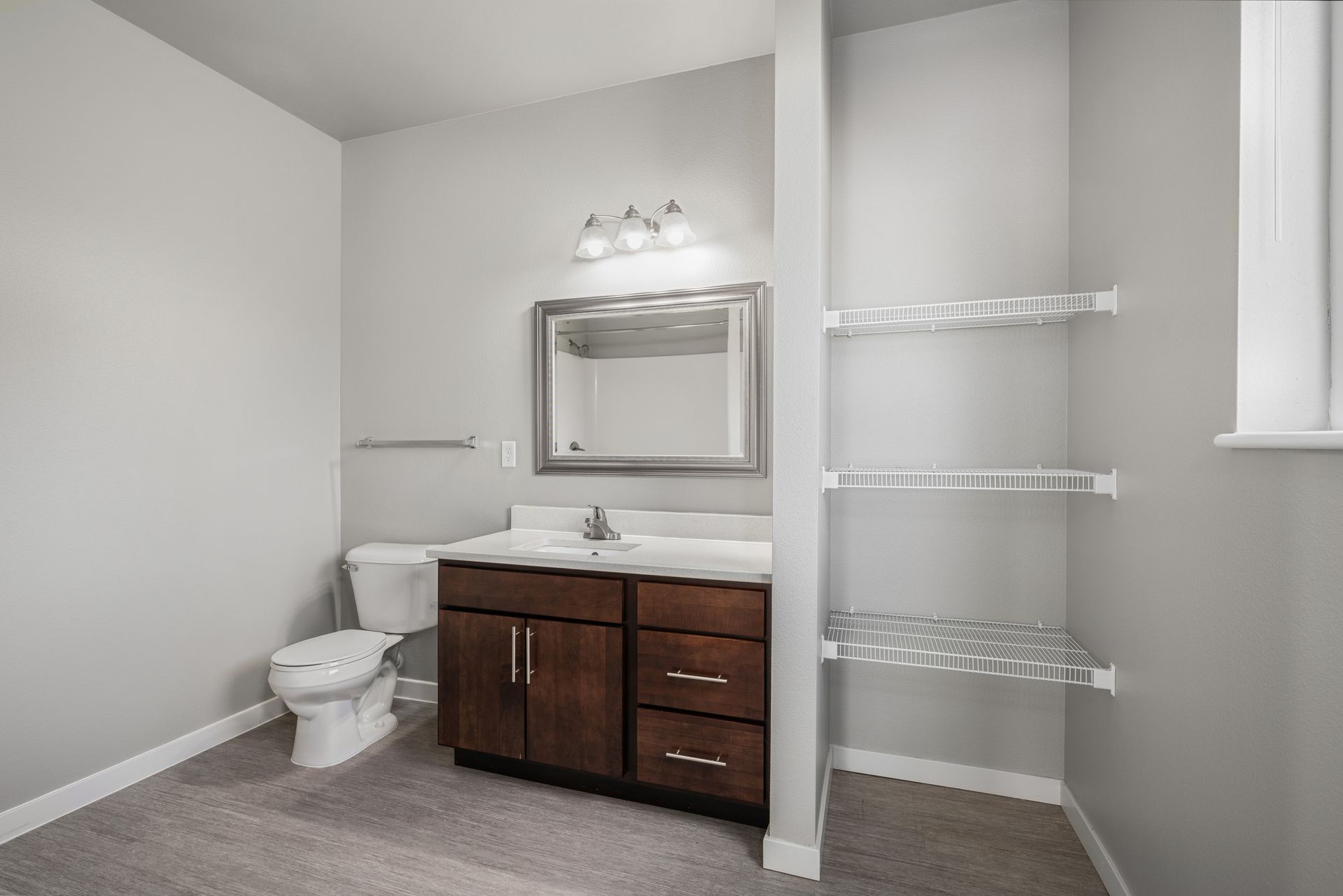 Bathroom with toilet, vanity, mirror, shelves, and gray walls.