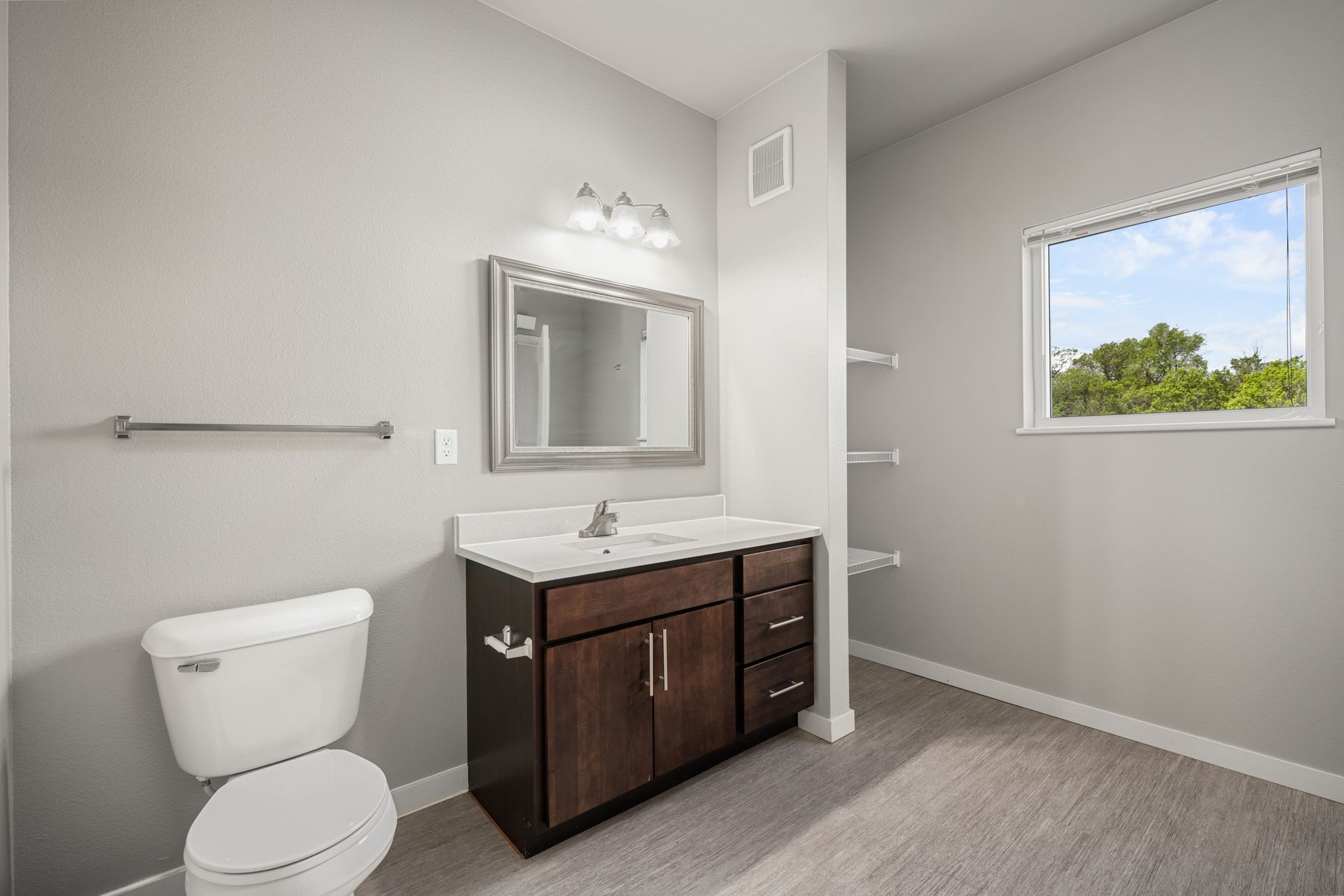 Bathroom with gray walls, dark brown vanity, toilet, mirror, and a small window with a view.