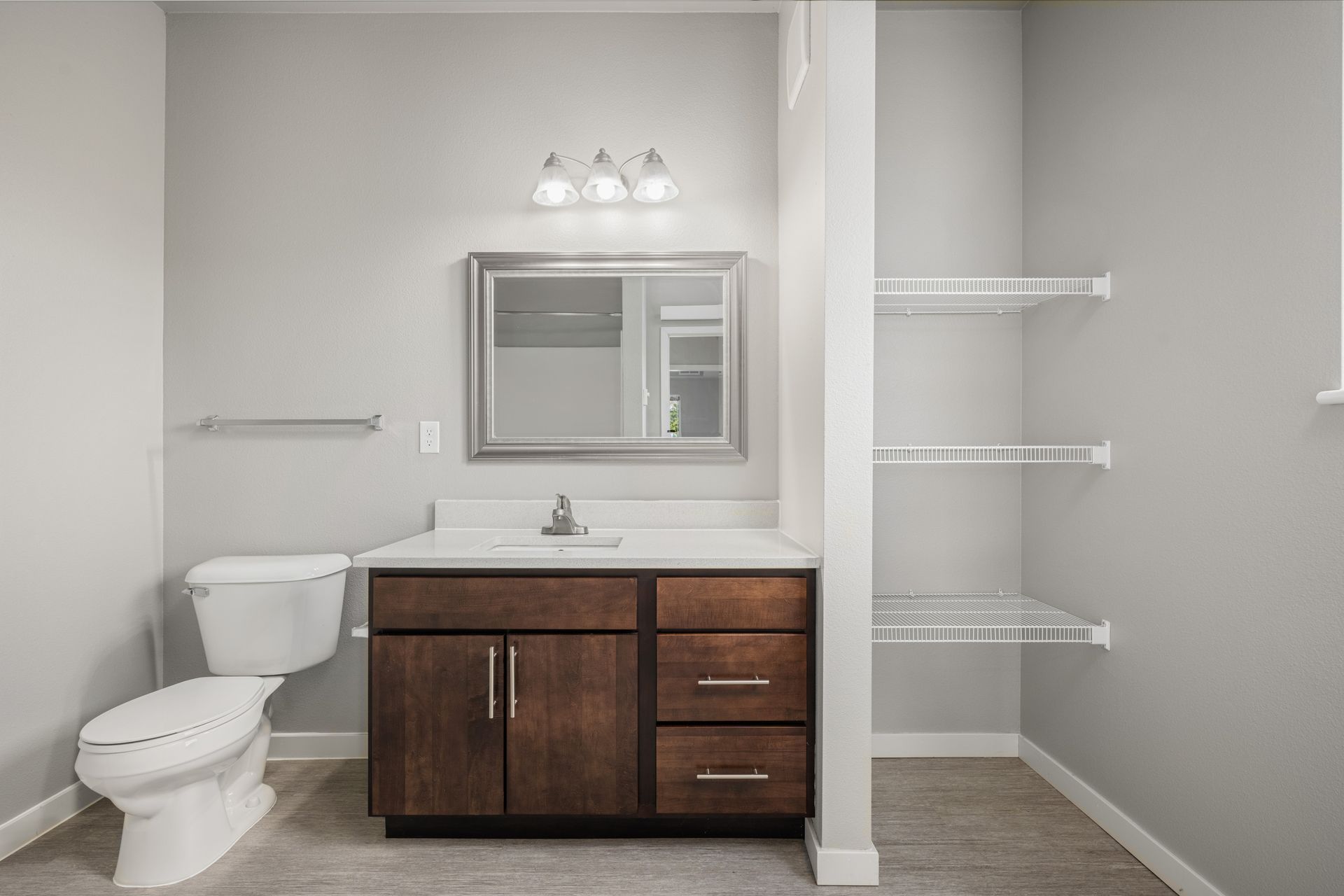 Bathroom with toilet, vanity, mirror, shelves, and gray walls.