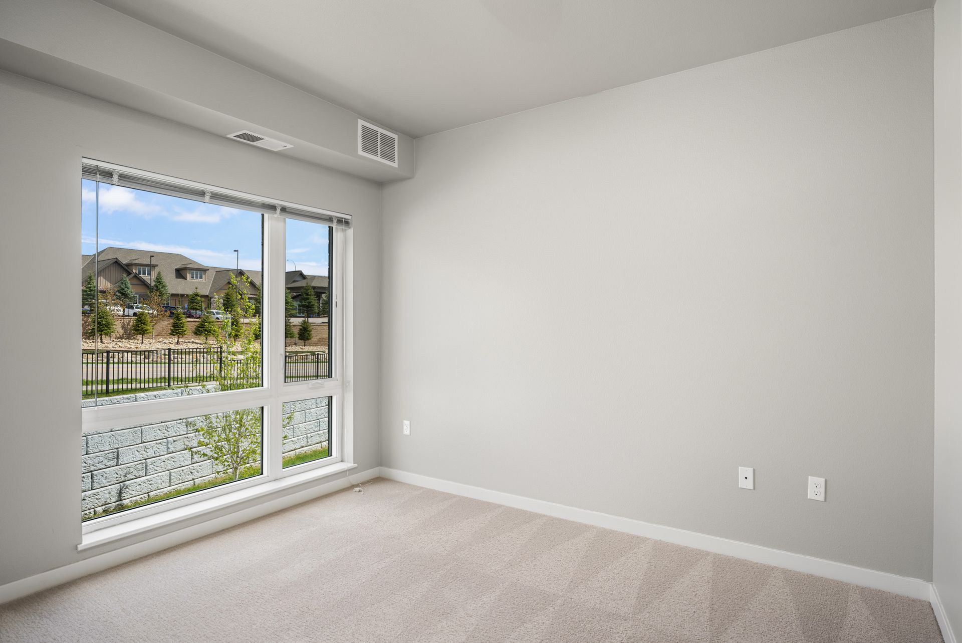 Empty room with large window overlooking a neighborhood and lake. Light gray walls, beige carpet.