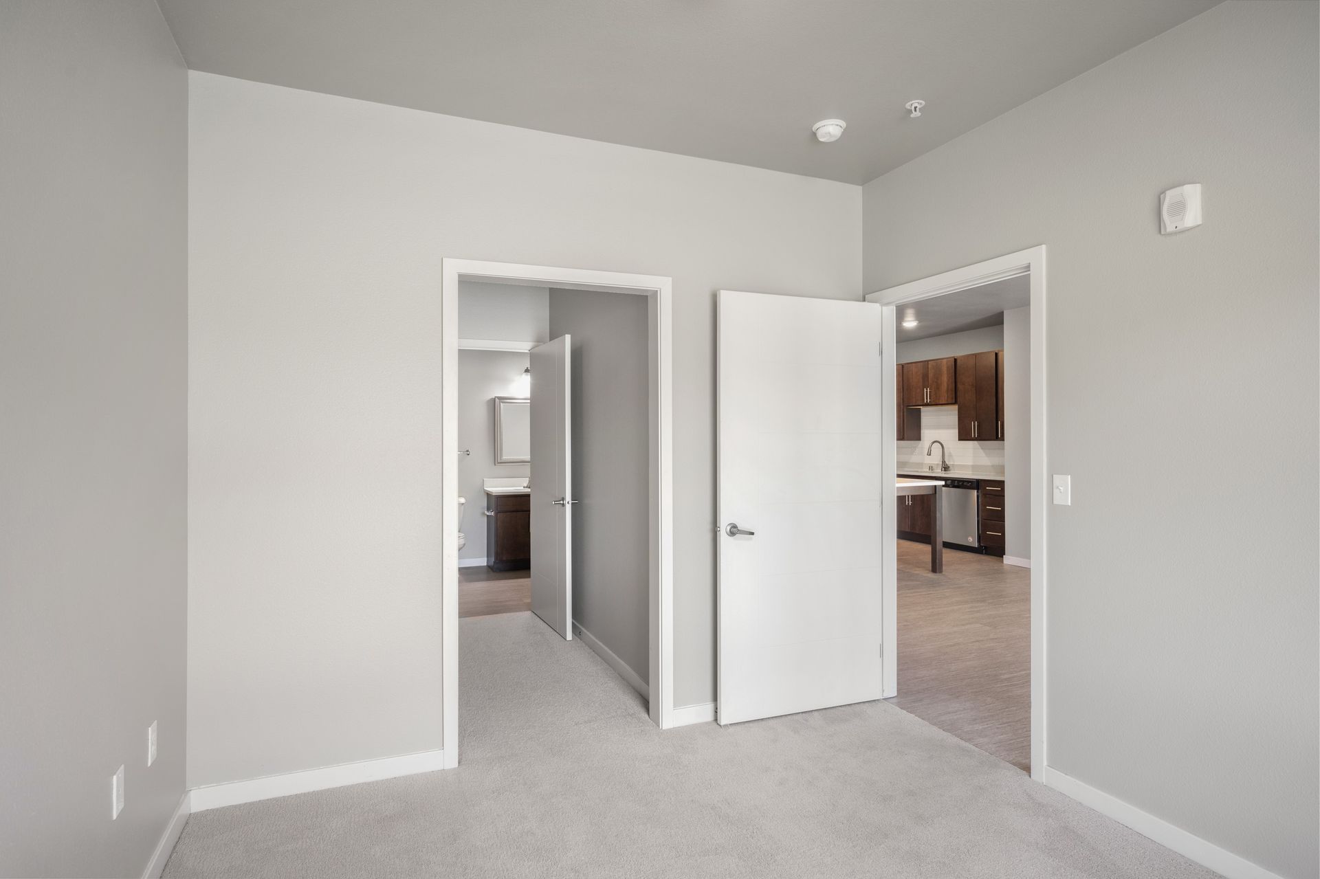 Empty room with carpet, two doorways leading to bathroom and kitchen.