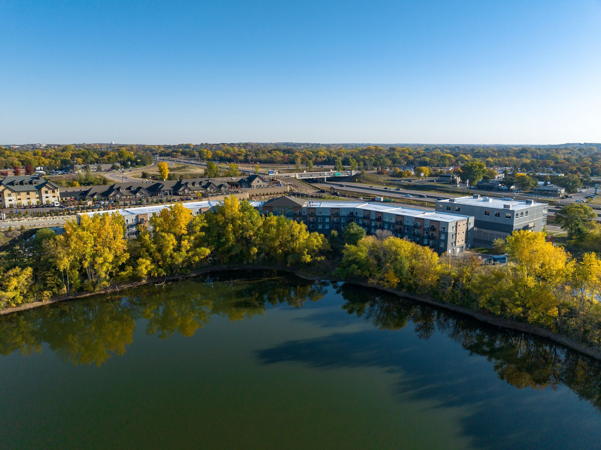 Aerial view of apartment complex beside a lake with trees displaying autumn colors; blue sky backdrop.