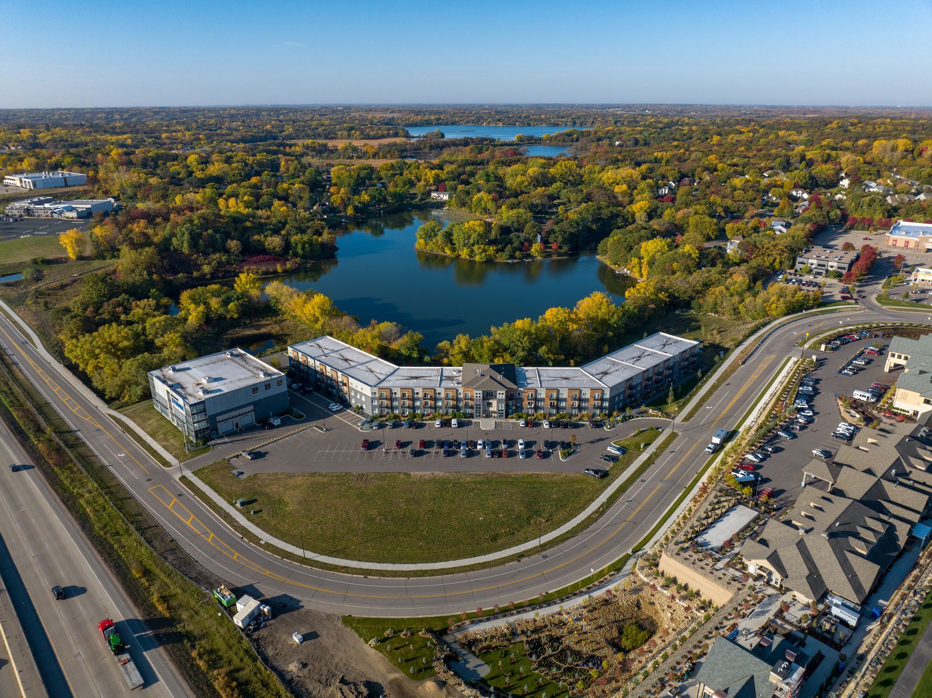 Aerial view: apartment complex and road curve near a lake surrounded by trees and a clear sky.