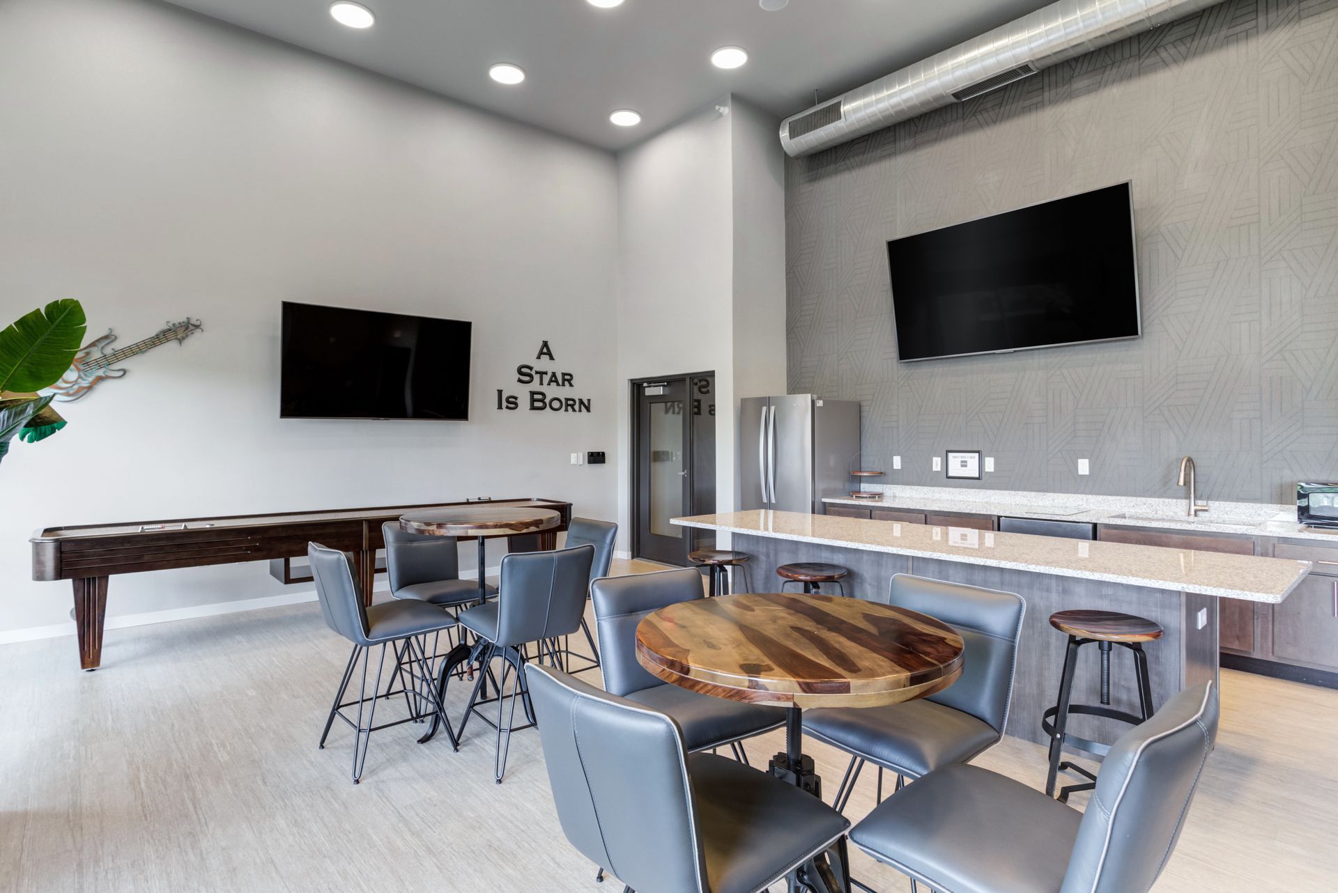 A modern lounge area with a shuffleboard table, bar, and TVs mounted on a gray wall.