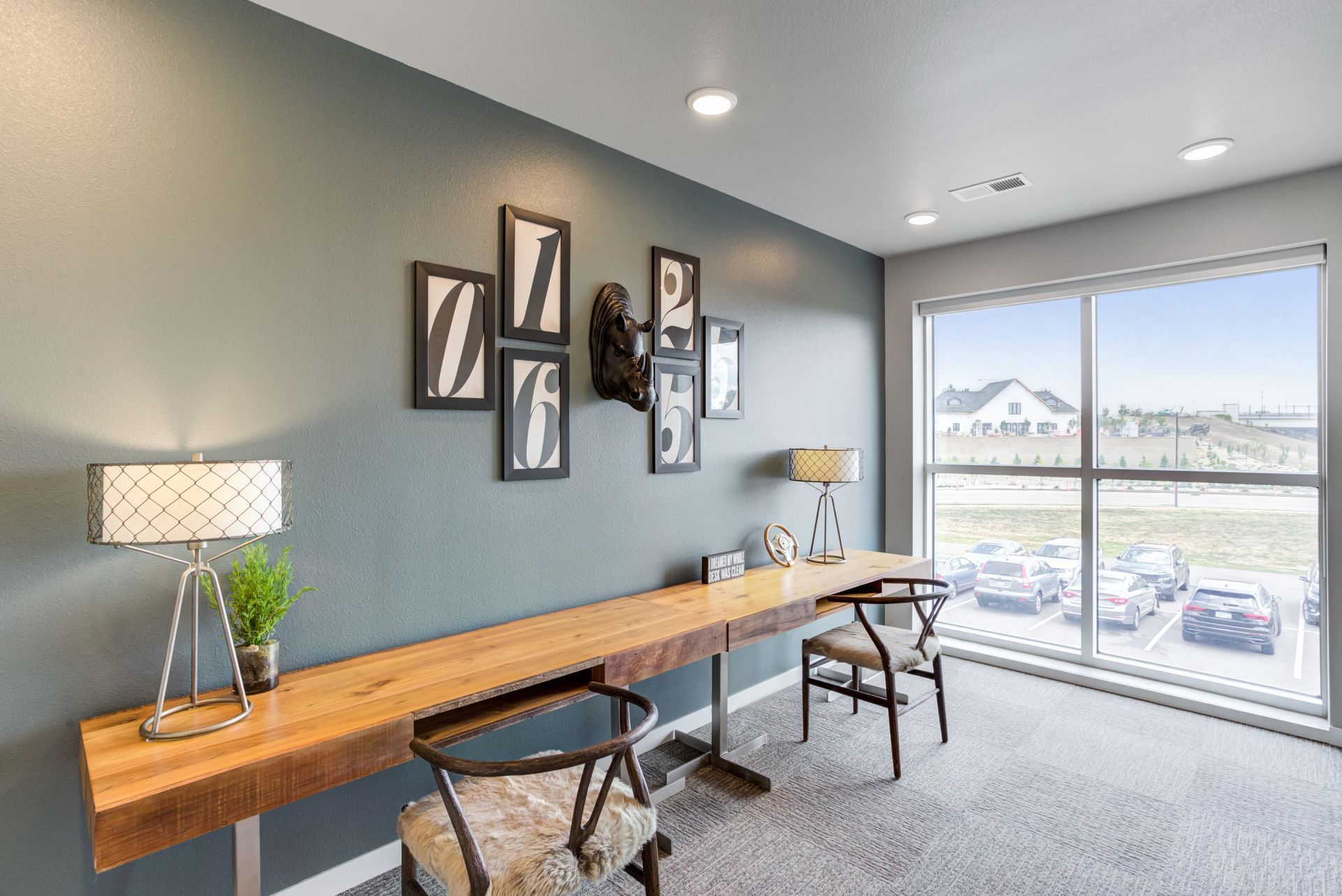 Desk with chairs, a window, and artwork on a blue-gray wall in a room with a window overlooking a parking lot.