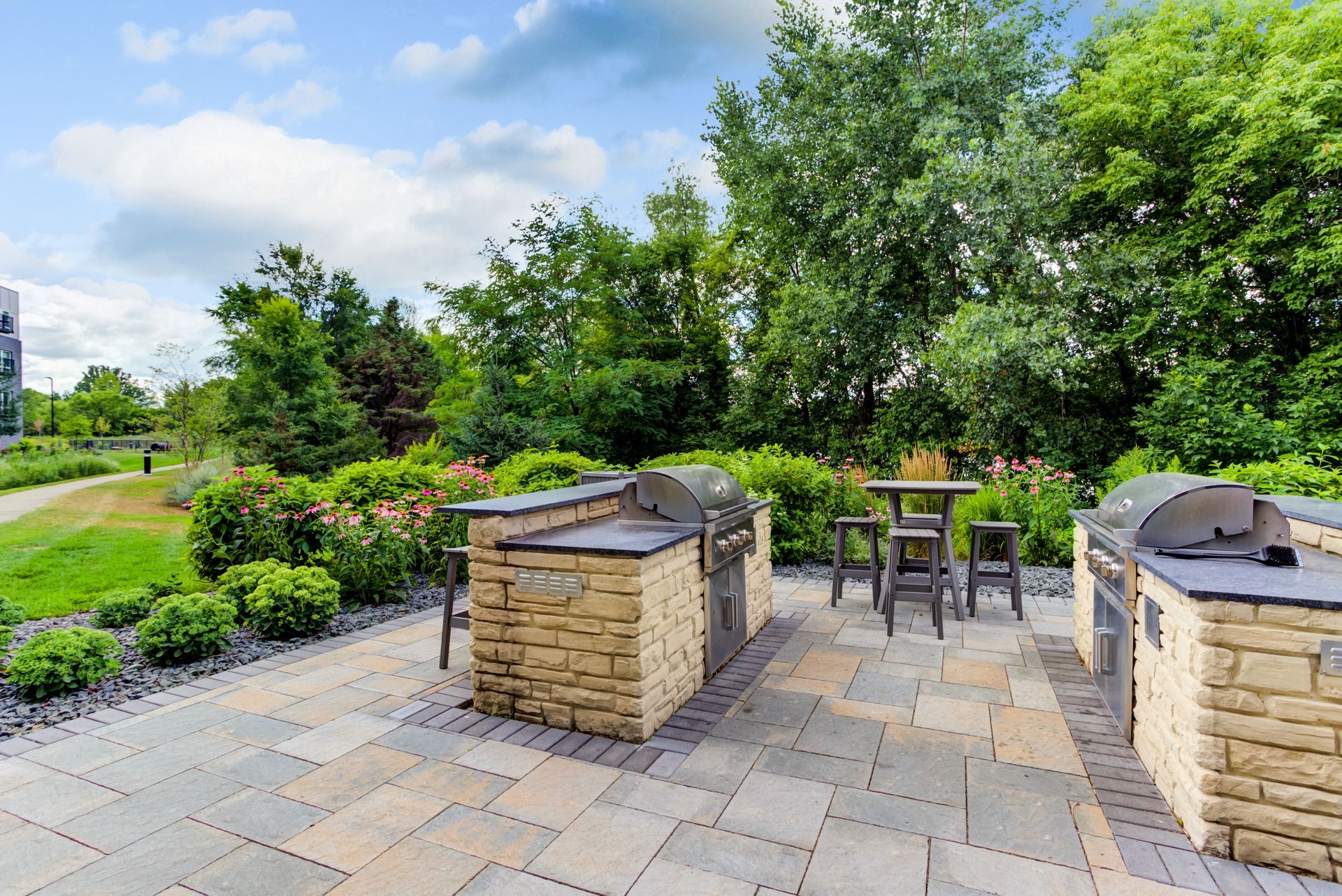Outdoor grilling area with stone brick structures, grills, and seating, surrounded by greenery and a paved patio.