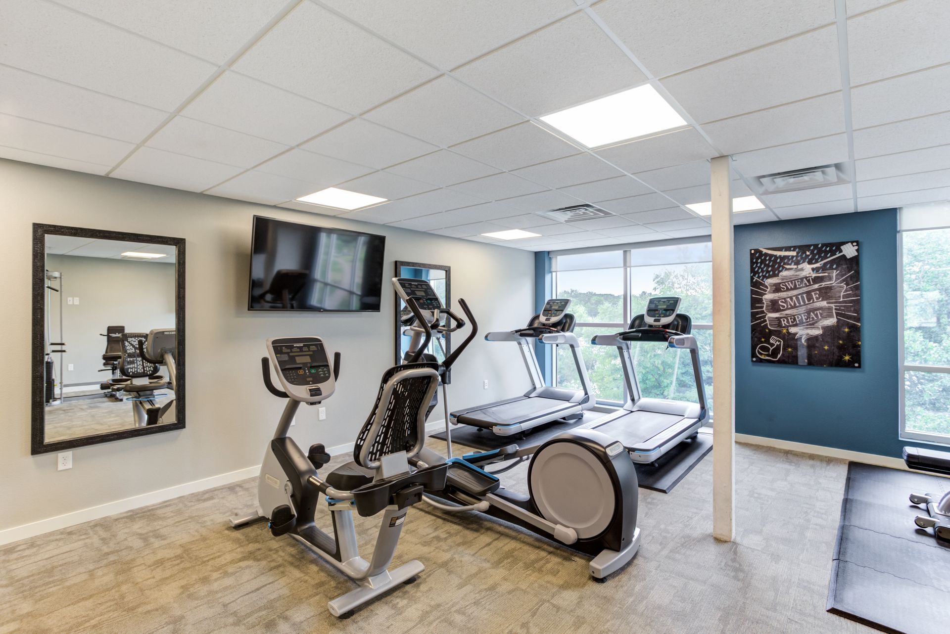 Gym with exercise equipment, including elliptical, treadmill, and recumbent bike, near large window with outdoor view.