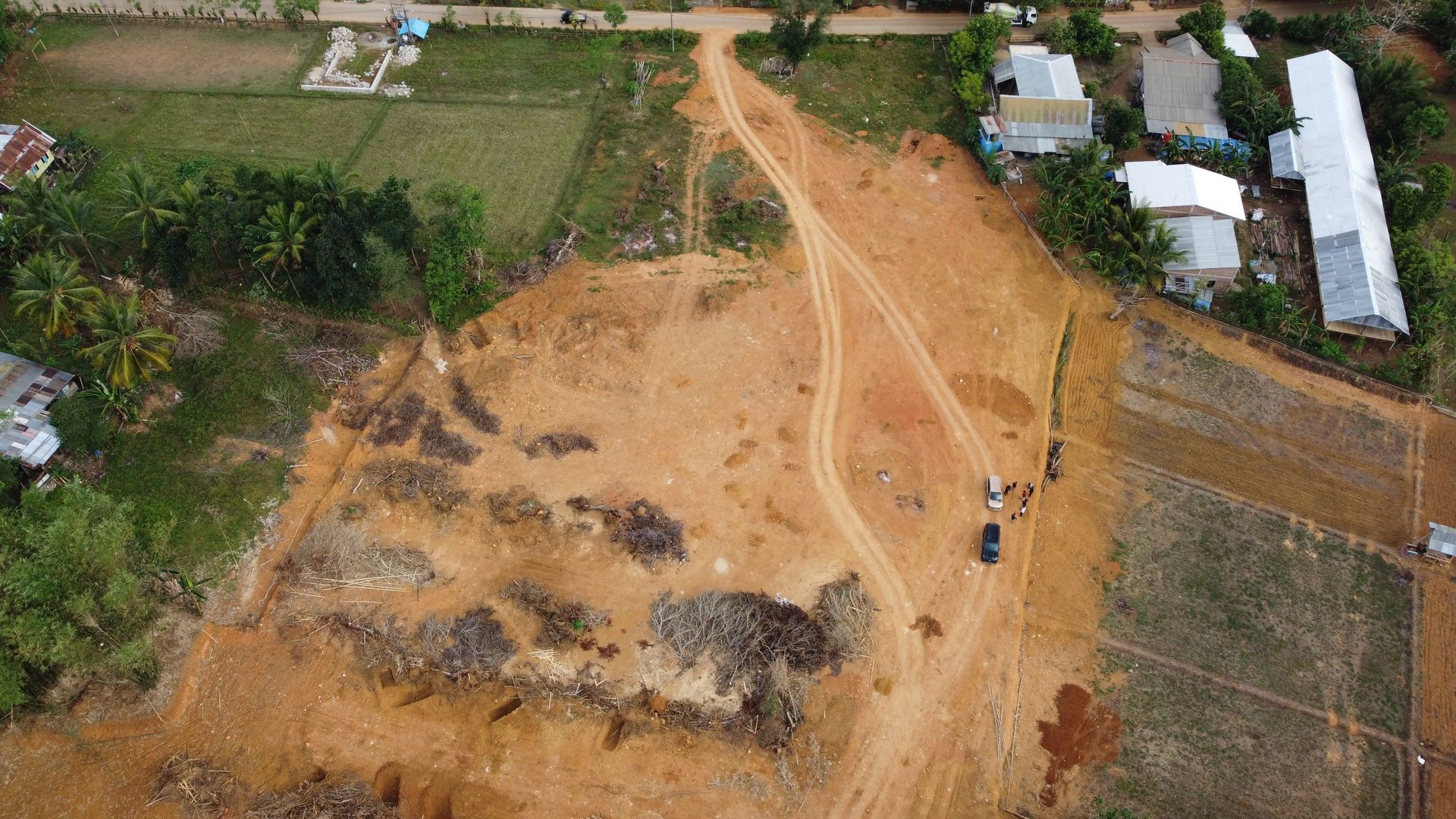 Aerial view of a cleared, reddish-brown land area with vehicle tracks, adjacent to green fields and buildings.
