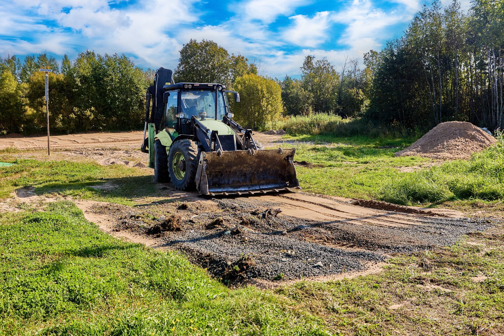 Backhoe leveling gravel in a grassy field, trees in the background, blue sky.