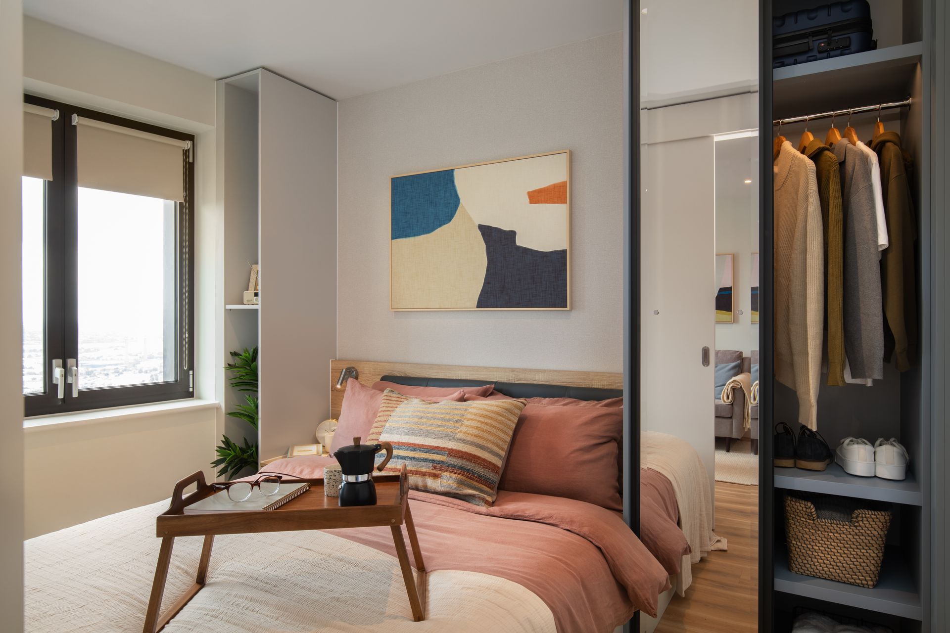A cozy bedroom with a bed topped with pink bedding and a breakfast tray, next to an open closet and a window.