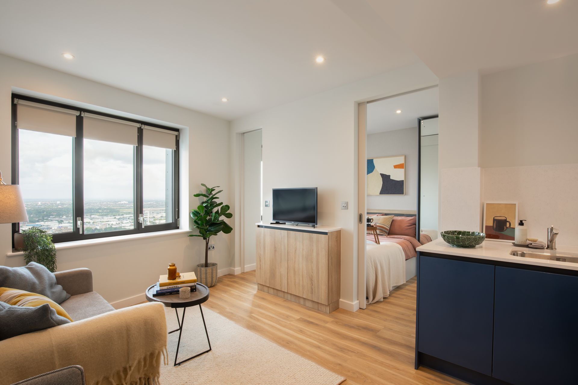 A modern, brightly lit apartment living area featuring a sofa, a small round table, a wooden TV stand, and an open bedroom.