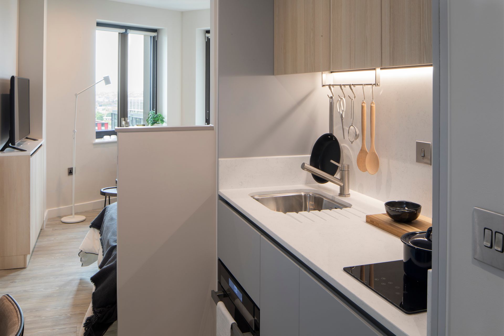 A modern, compact kitchenette with white counters and light wood cabinets, opening to a view of a tidy bedroom area.