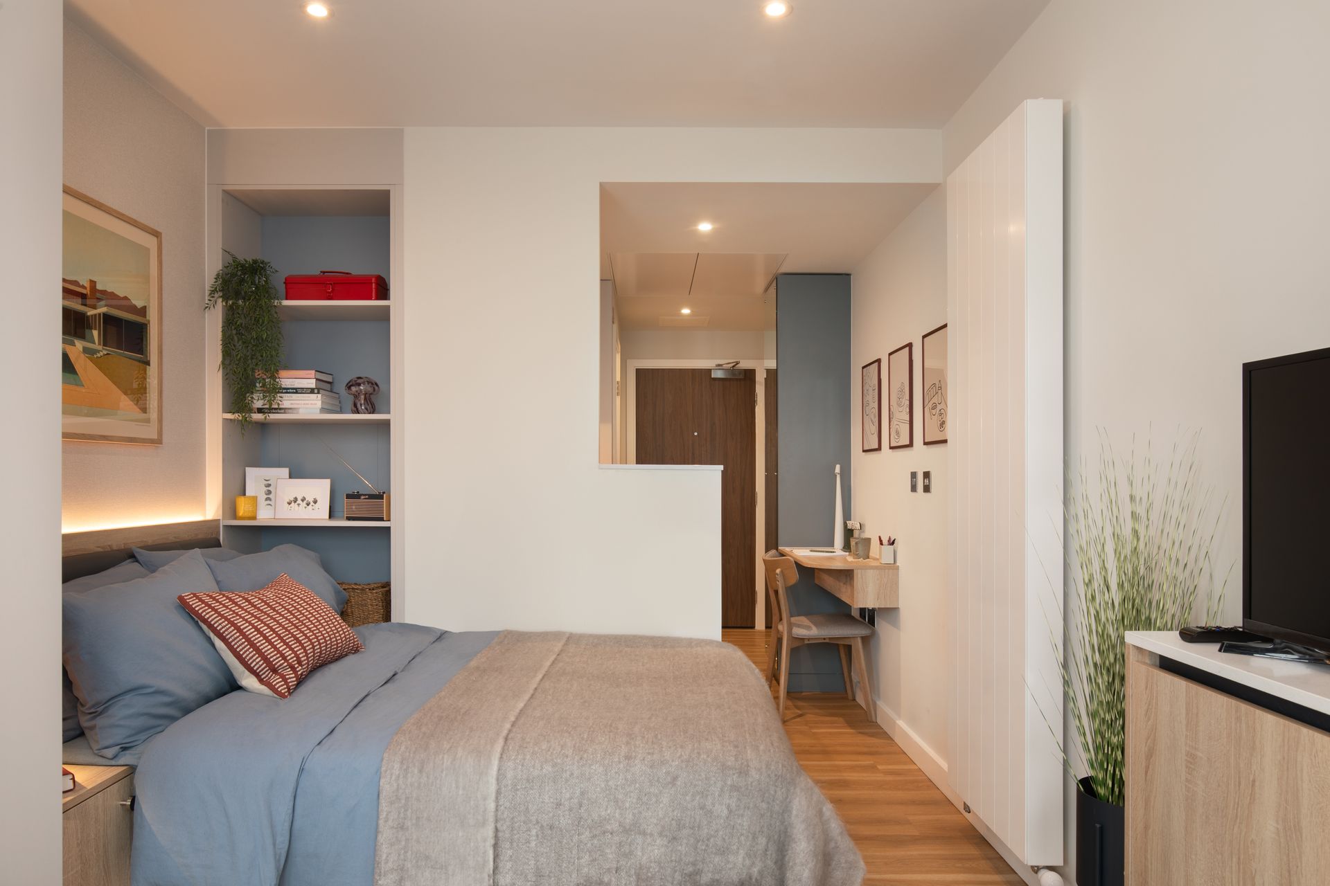 A compact bedroom featuring a blue-toned bed, adjacent built-in shelving, a small desk area, and light wood flooring.