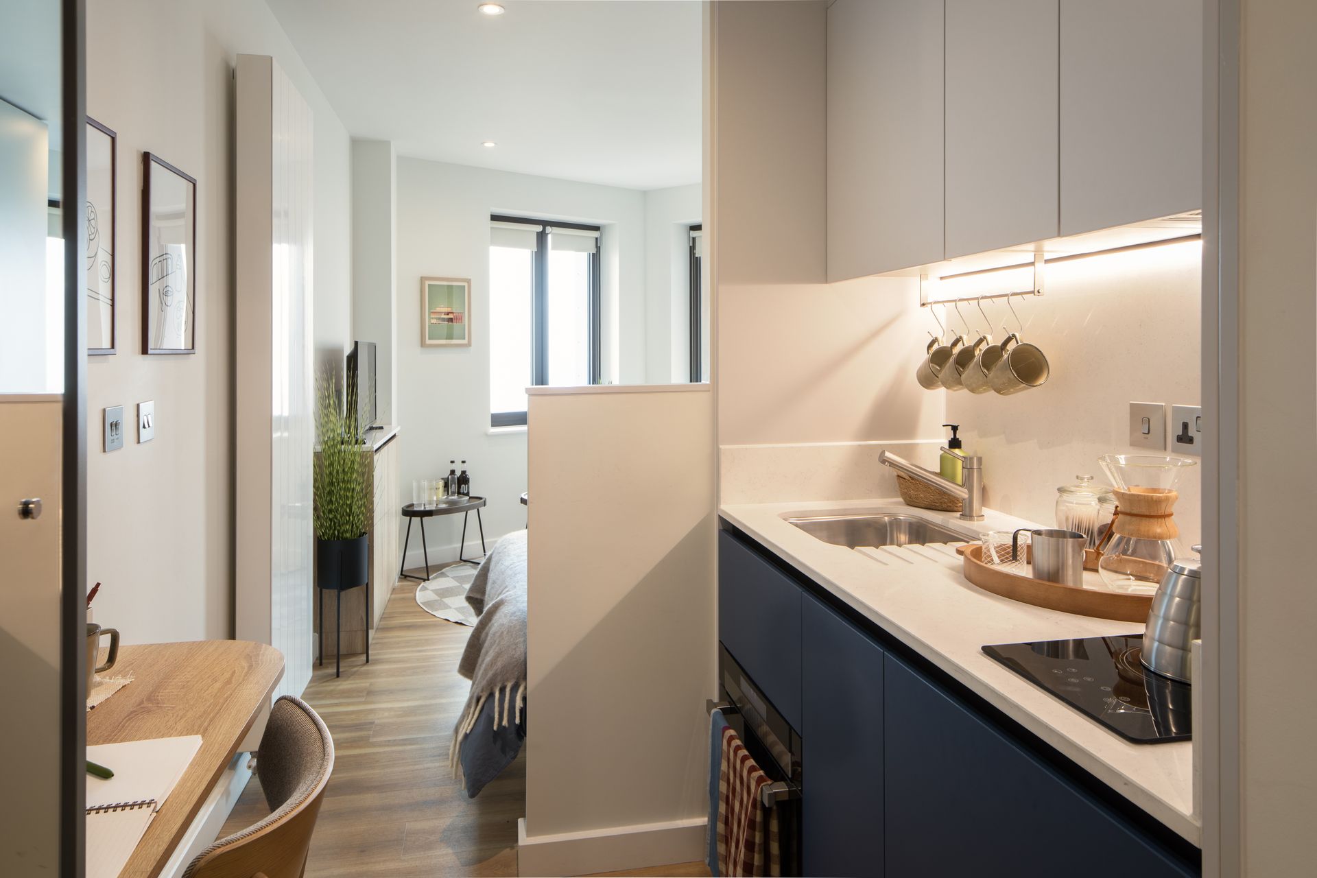 A compact studio apartment features a kitchenette with dark blue cabinets, a light wooden desk, and a bedroom area.