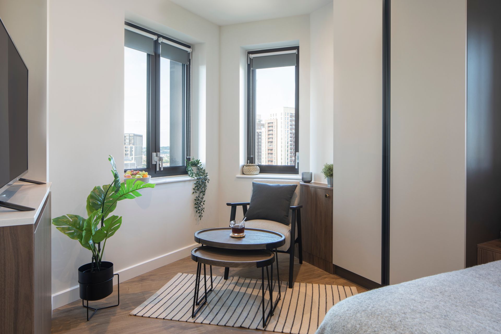 A cozy corner with a chair, nested tables, a potted plant, and windows showing a city view.