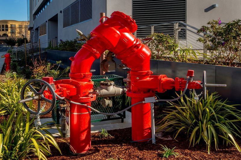 Hand holding HVAC gauges connected to copper pipes; red, blue, and yellow hoses against a brick wall. Red fire hydrant and pipes in front of a building, surrounded by greenery.