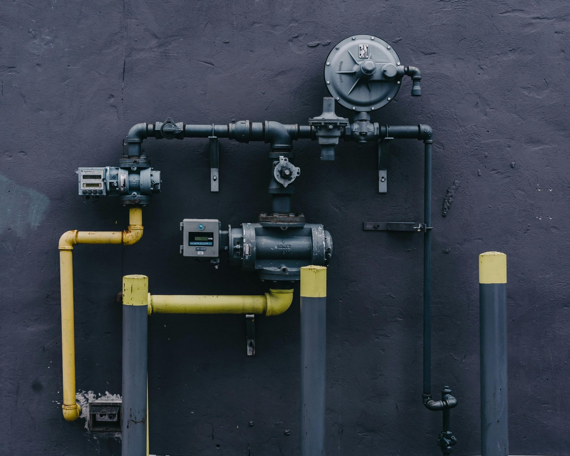 Hand holding HVAC gauges connected to copper pipes; red, blue, and yellow hoses against a brick wall. Red fire hydrant and pipes in front of a building, surrounded by greenery.