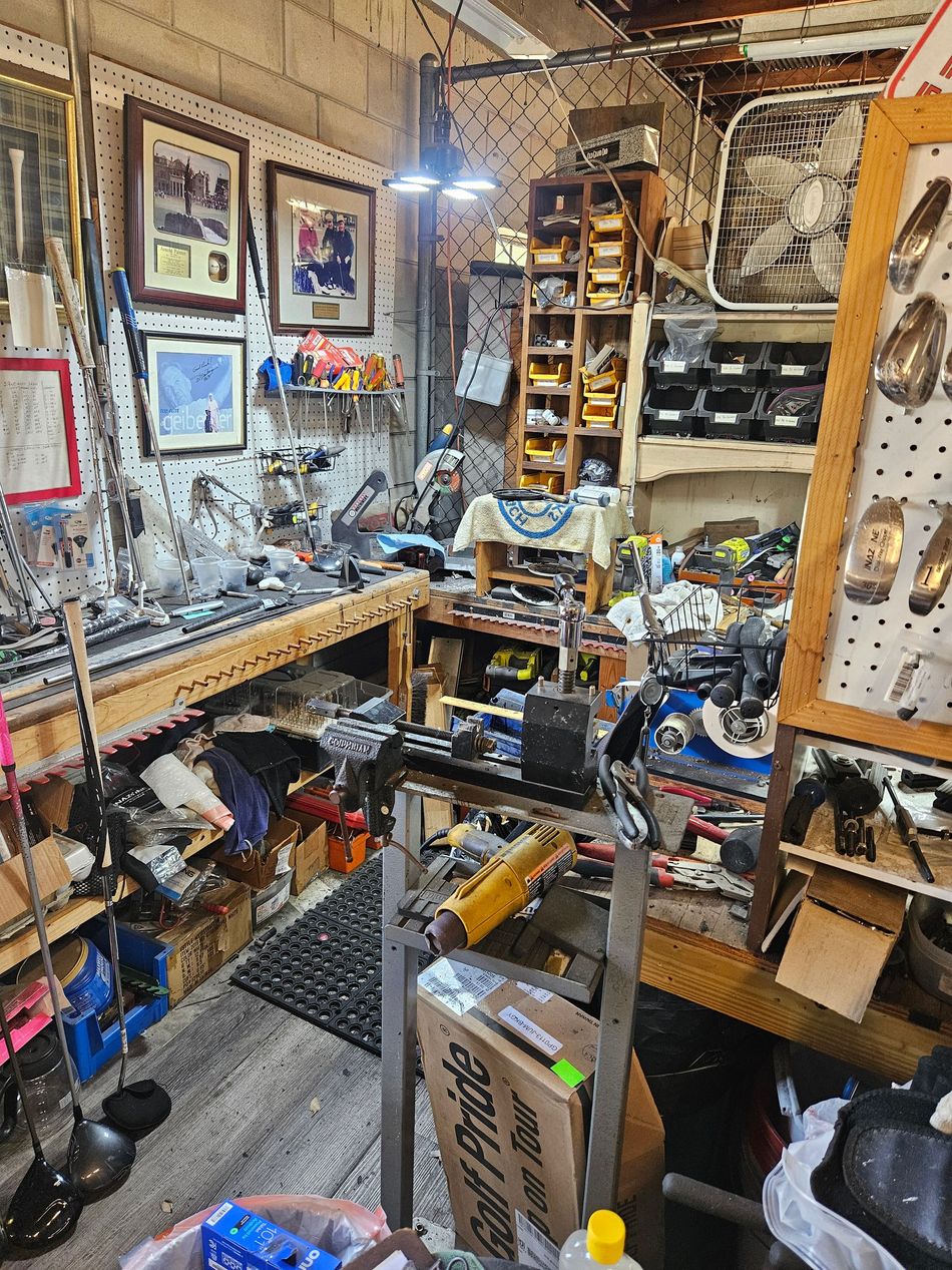 Cluttered workshop with shelves, tools, framed pictures, and a narrow aisle under warm lighting