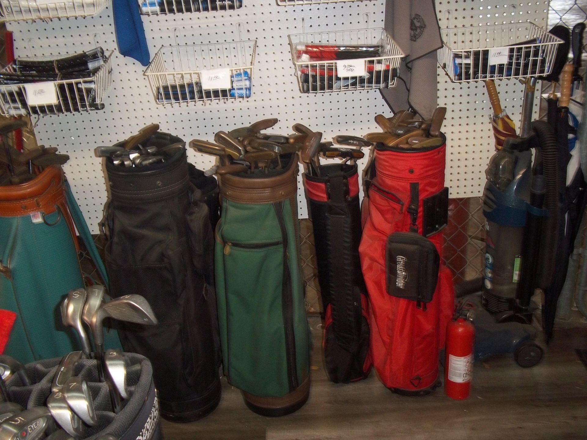 Several golf bags in black, green, and red stand against a wall with wire baskets, alongside a red fire extinguisher.