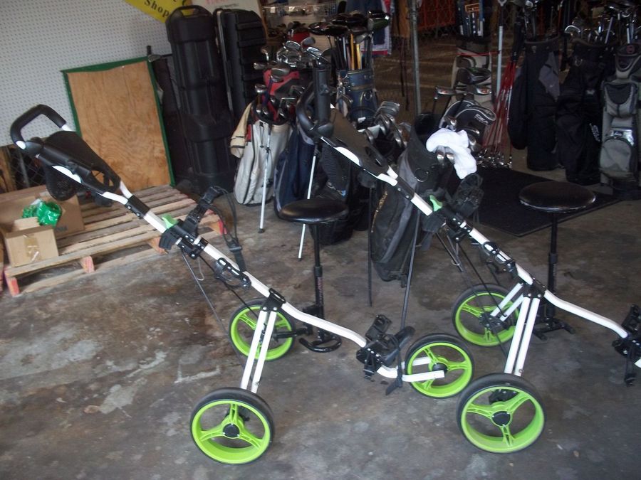 Two white golf push carts with neon green wheels parked indoors, with golf bags in the background.
