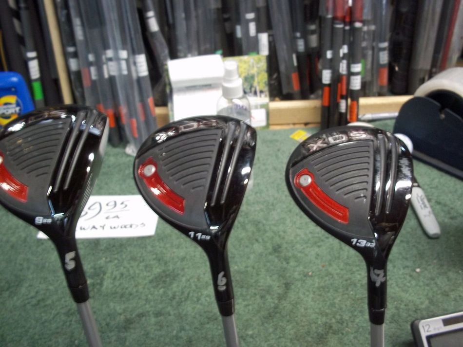 Three black golf fairway woods—labeled 5, 6, and 7—with red accents on the heads, standing upright on a green surface.