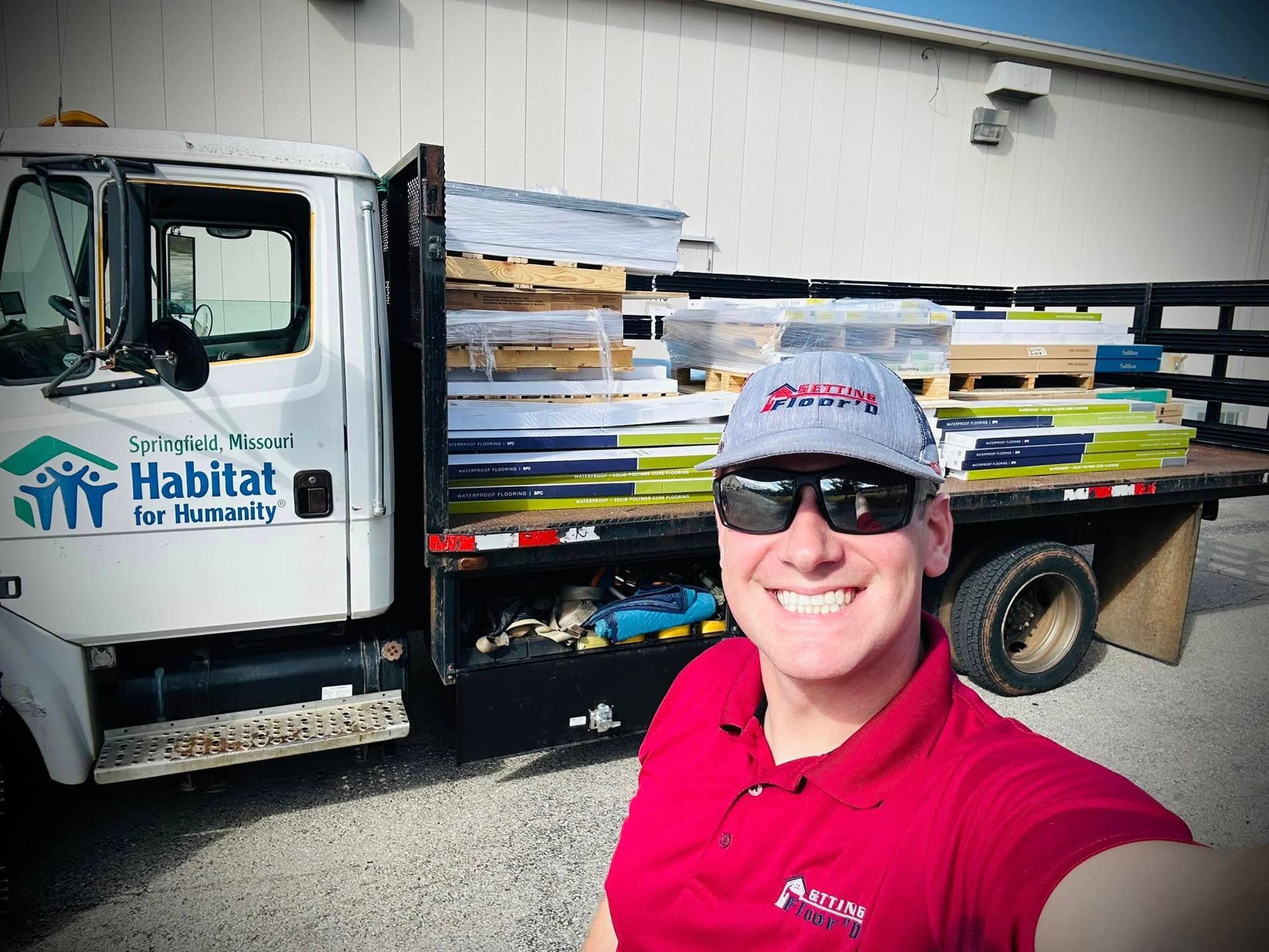 Man in red shirt and cap smiles next to Habitat for Humanity truck loaded with building materials.