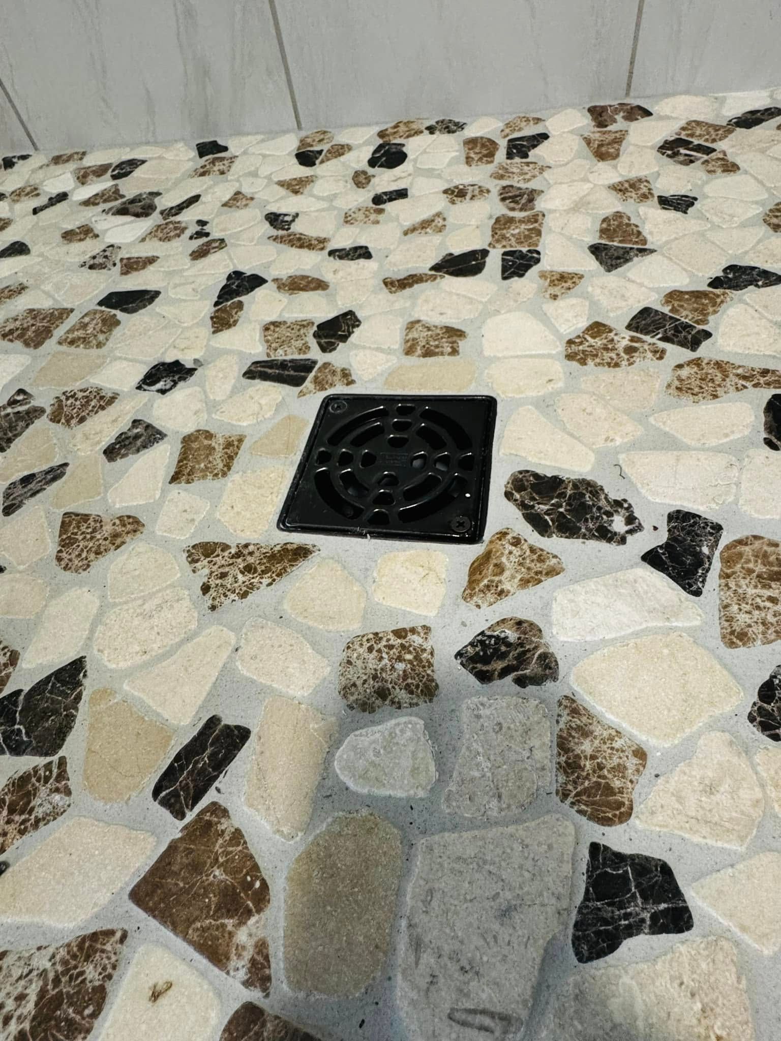 Shower floor with black drain, featuring irregularly shaped beige, brown, and black stones.