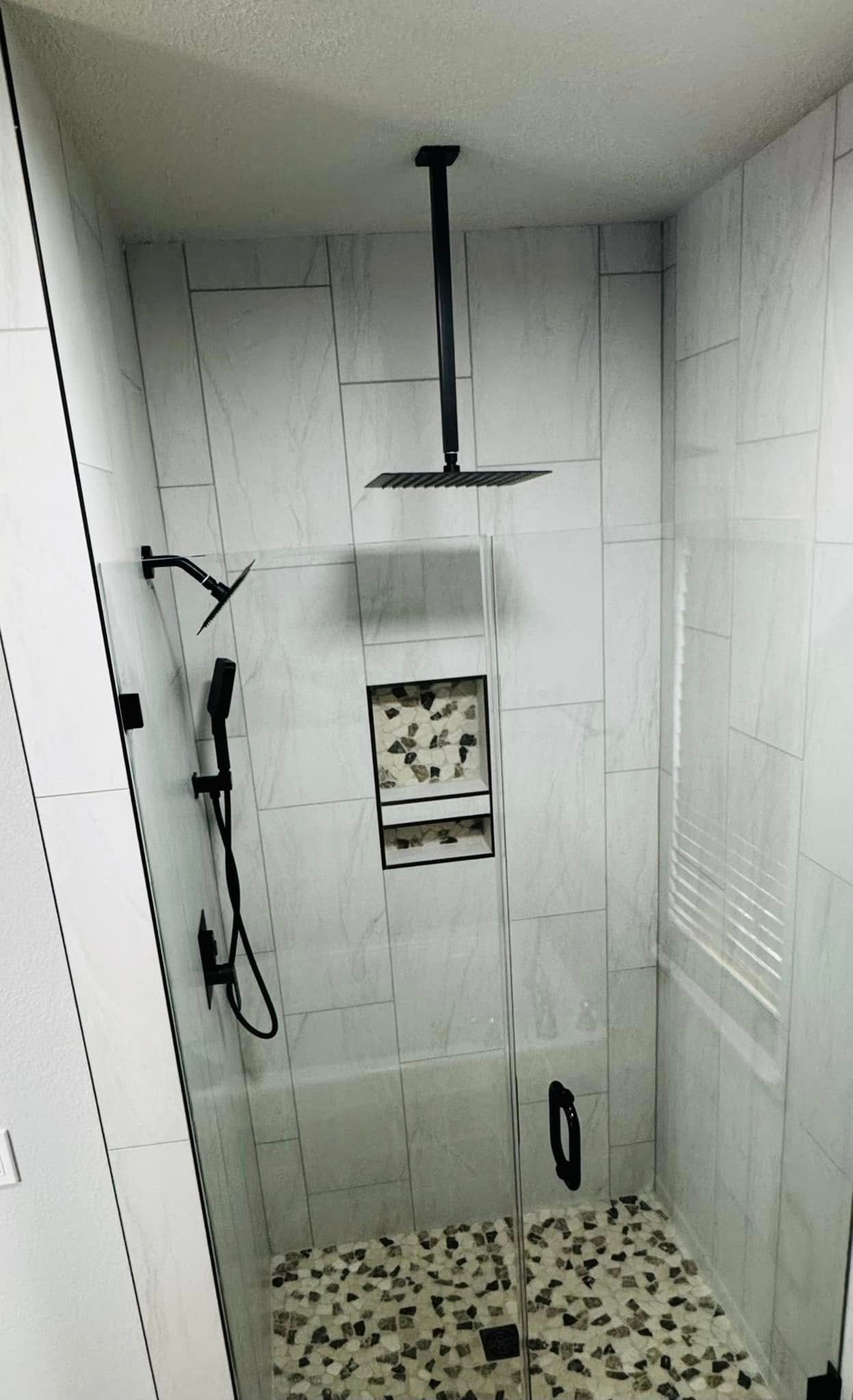 Modern shower with black fixtures, white tiles, glass door, and pebble floor.