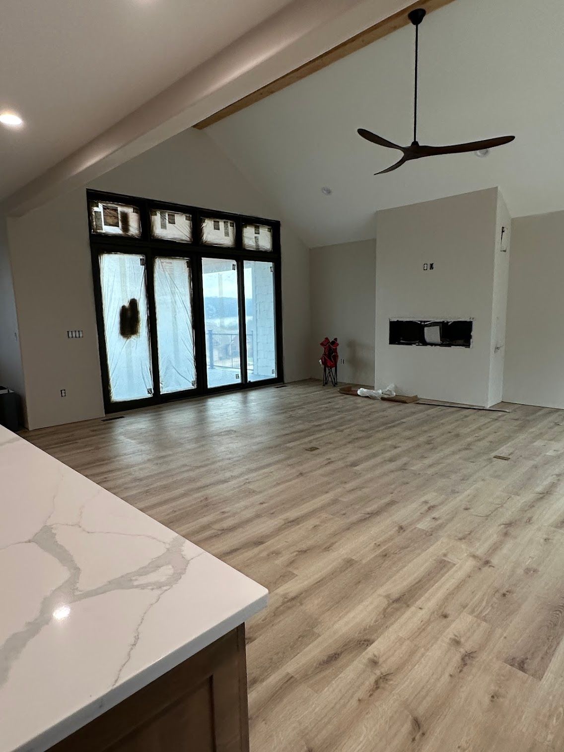 Open-concept living space with light wood flooring, large windows, a fireplace, and a dark ceiling fan.