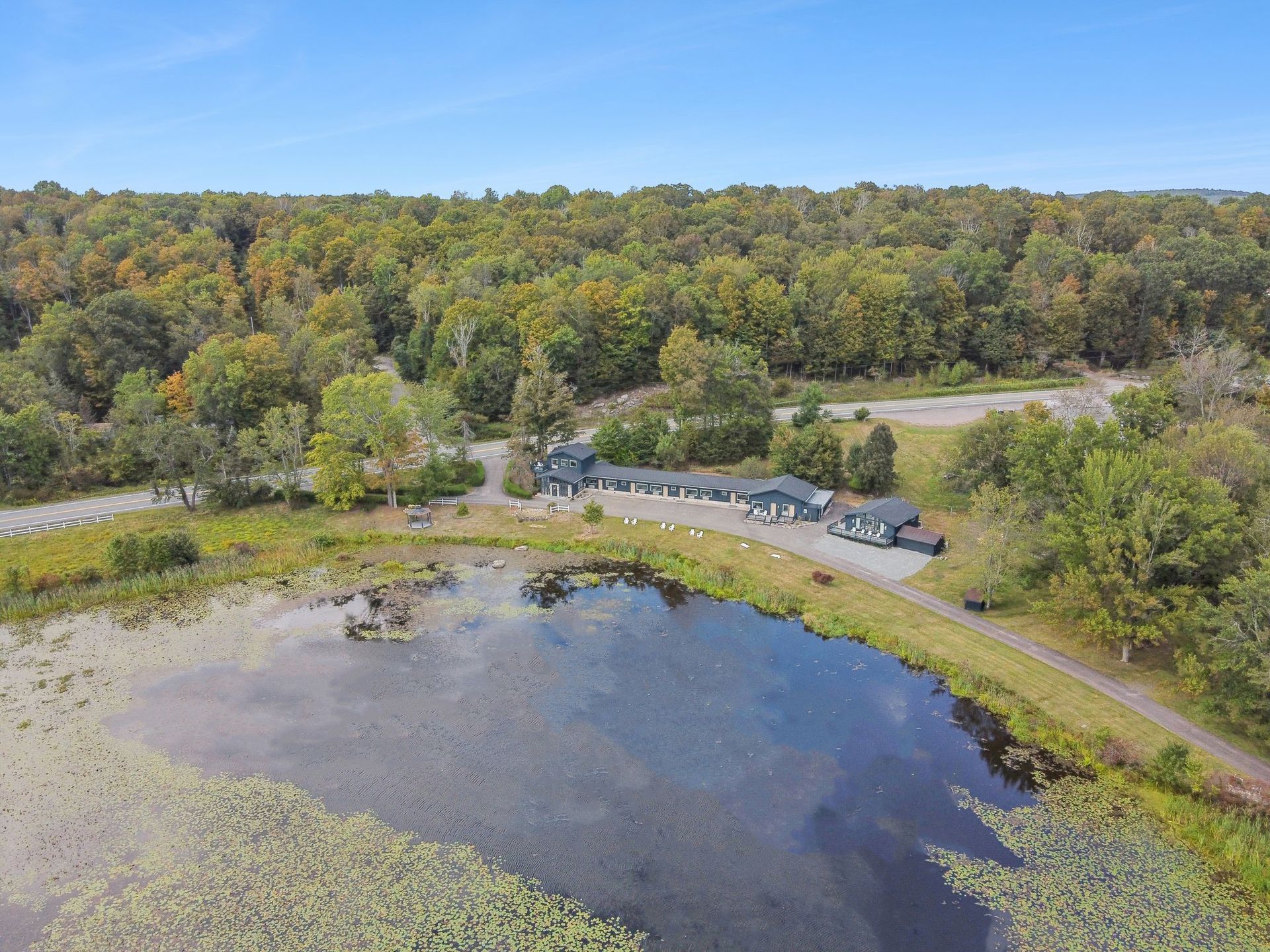 Aerial view: building complex near a pond with lily pads, surrounded by trees under a blue sky.