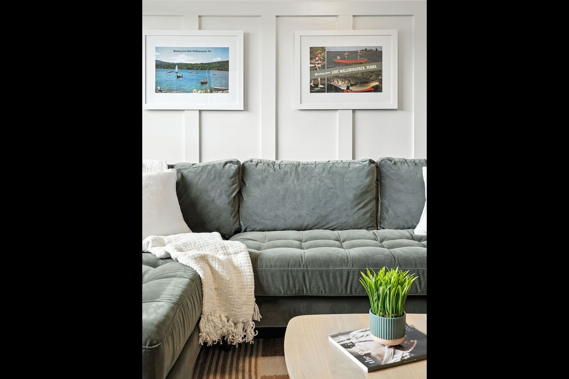 A living room with a gray couch, white wall, and framed art. A blanket and plant sit on the coffee table.